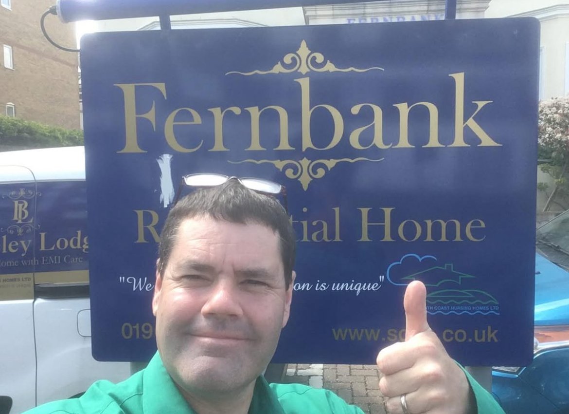 A man wearing a green shirt giving a thumbs-up in front of a large blue sign for Fernbank Residential Home. The sign includes the home's name, a quote, and contact information.