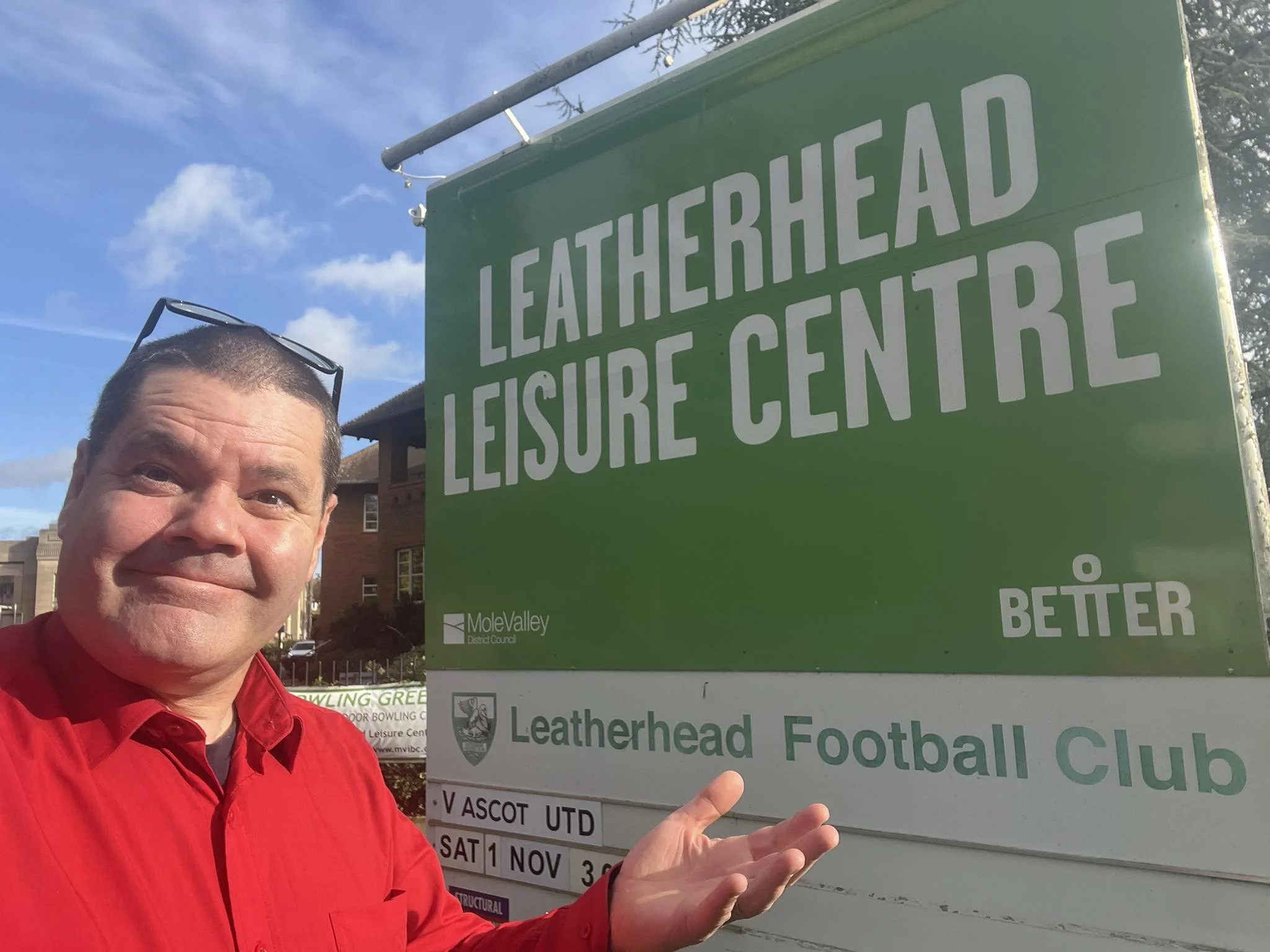 A man in a red shirt standing next to a green sign for Leatherhead Leisure Centre, pointing towards the sign and smiling. The sky is blue with some clouds, and there are buildings in the background.