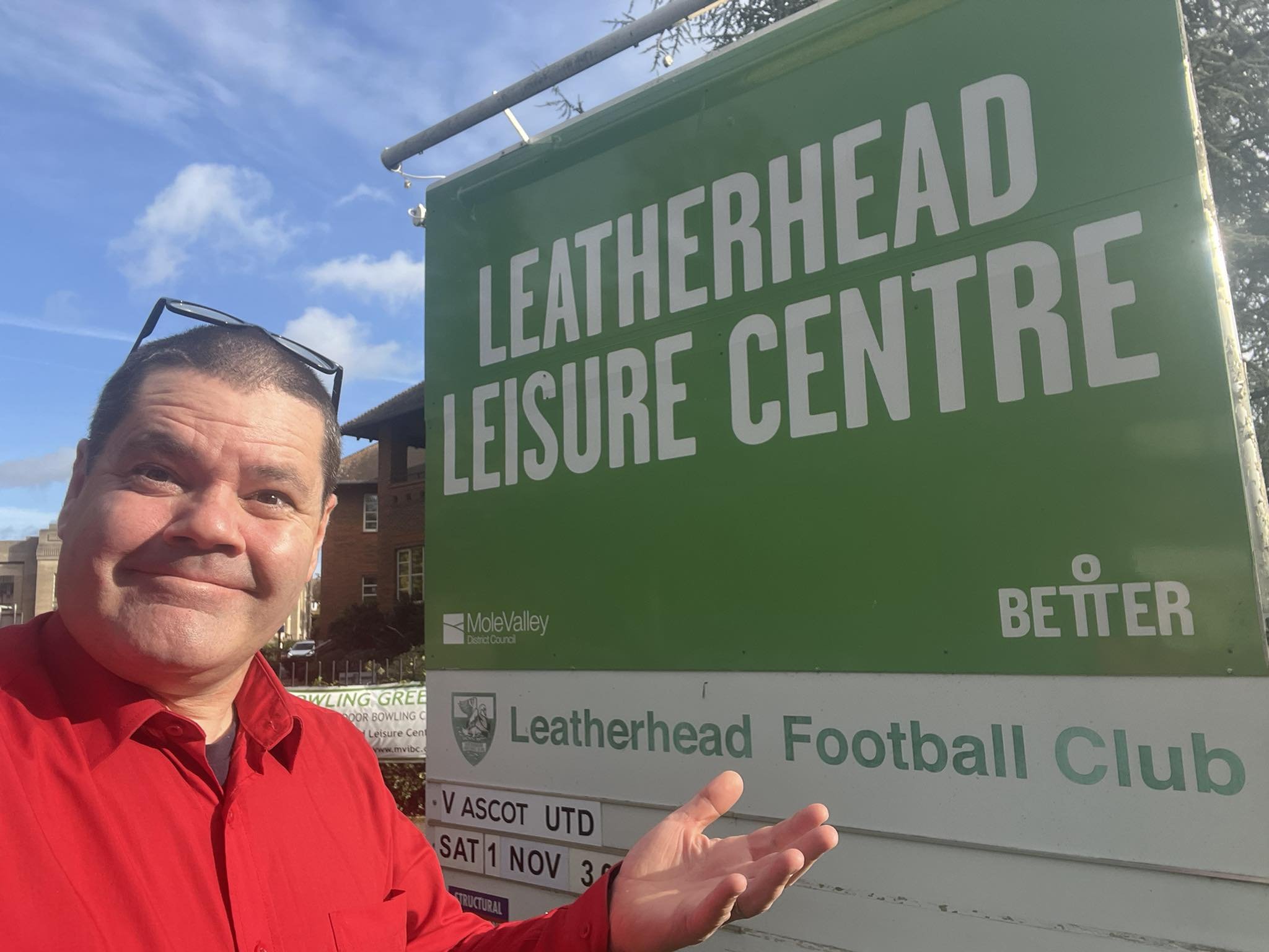 A man in a red shirt standing next to a green sign for Leatherhead Leisure Centre, pointing towards the sign and smiling. The sky is blue with some clouds, and there are buildings in the background.
