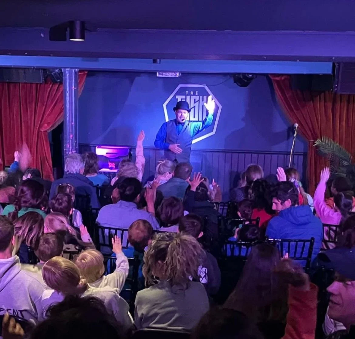 A performer on stage at a comedy club with a dark background and red curtains, engaging with an audience of young people, some raising their hands.