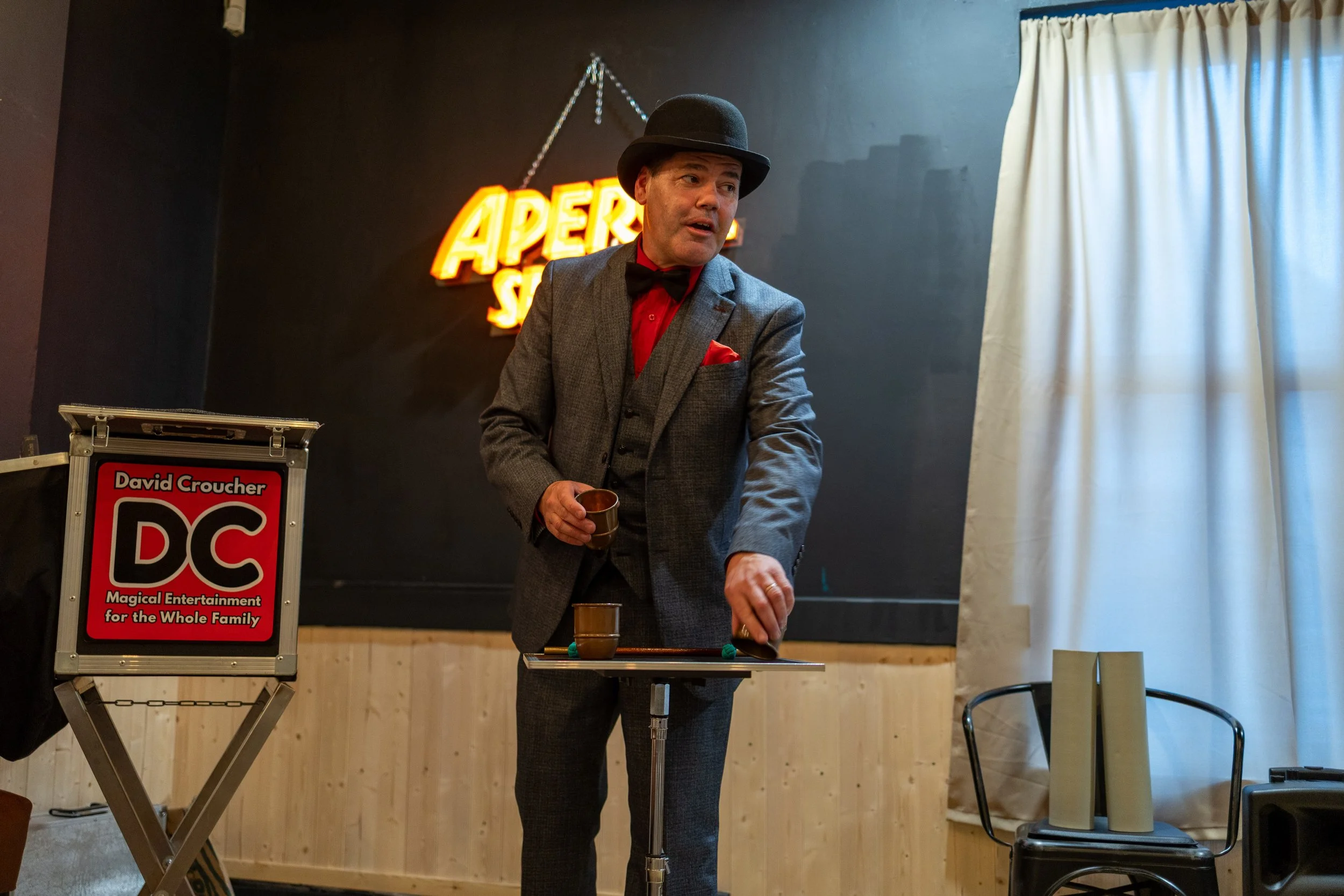 A man dressed in a gray suit, red shirt, black bow tie, and black bowler hat performing a magic trick on stage with a table and cups. Behind him is a neon sign that reads 'Aperture', and a wooden-paneled wall with a white curtain on the right side.