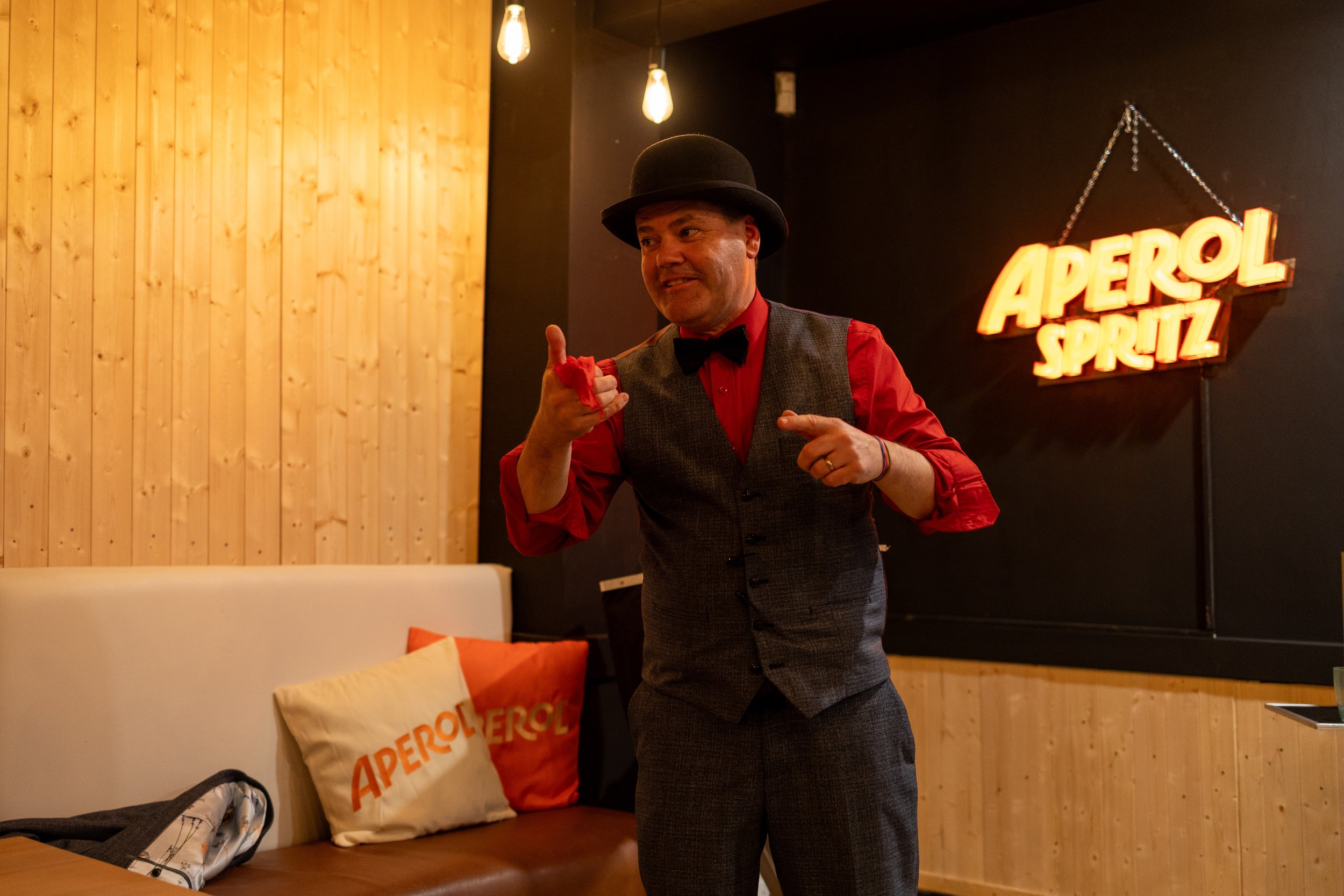 A man dressed in a red shirt, gray vest, black bow tie, and black bowler hat pointing and smiling in a cozy, warmly lit room with wood-paneled walls and an illuminated