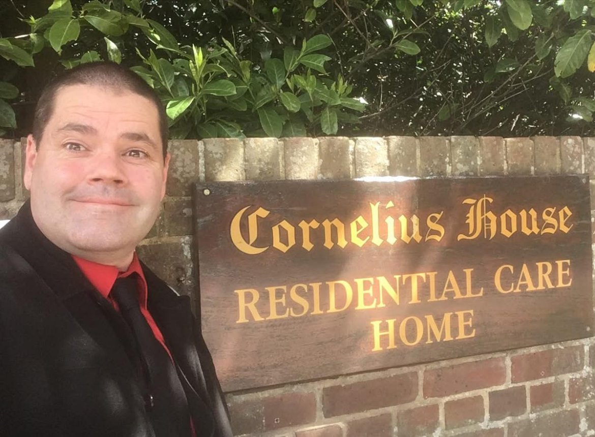 A man in a black jacket with a red shirt taking a selfie in front of a sign that reads "Cornelius House Residential Care Home" with green foliage and a brick wall in the background.