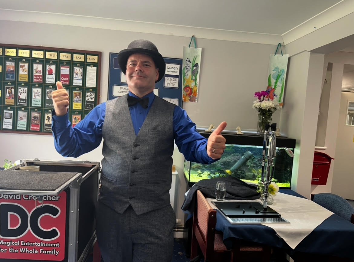 A man dressed in a vintage style, wearing a bowler hat, a blue shirt, a gray vest, and a bow tie, standing in a room with an aquarium, colorful decorations, and a table with a glass of water, giving a thumbs-up gesture.