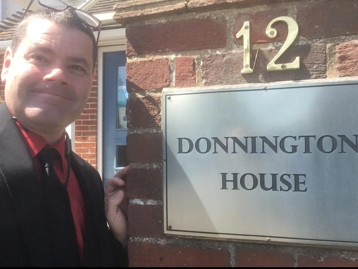 A man in a suit and red shirt stands outside a brick building next to a sign that reads "DONNINGTON HOUSE" with the house number 12 above it.