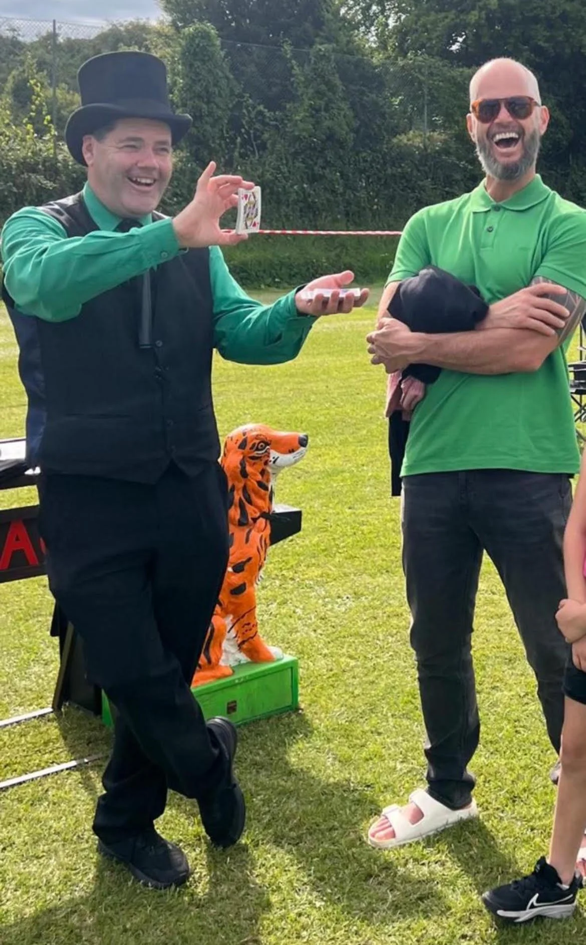 Two men laughing outdoors, one dressed as a magician with a top hat and a bright green shirt, performing a trick with a playing card and string. A tiger puppet is on a green box nearby.