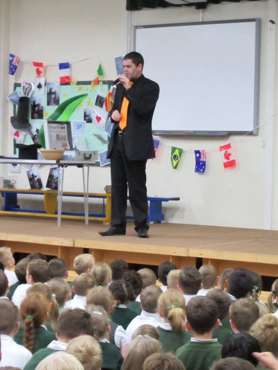 A man in a black suit and orange tie speaking into a microphone on stage, with a crowd of children seated in front. The stage is decorated with international flags, photos, and a whiteboard.