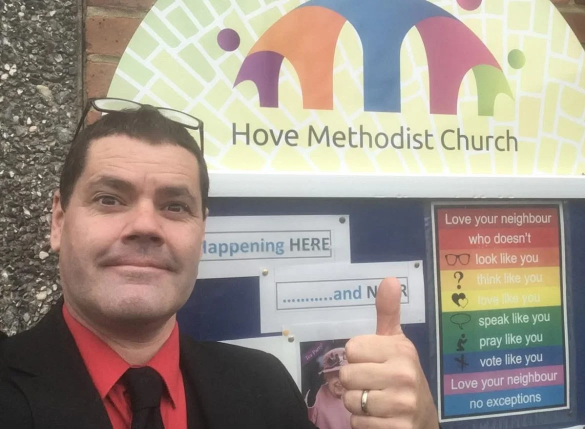 A man wearing a suit and red shirt giving a thumbs up in front of a community notice board and a church sign that says "Hove Methodist Church." The notice board has colorful sections with messages advocating love, kindness, and respect for neighbors.