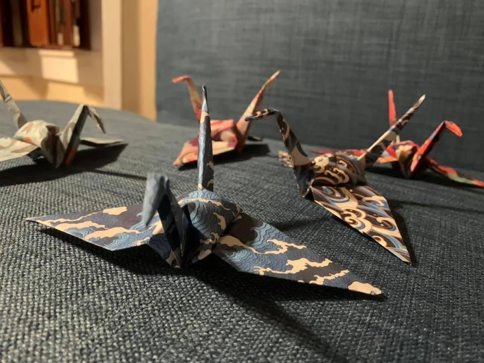 Multiple origami paper cranes with colorful patterned paper on a dark fabric surface.