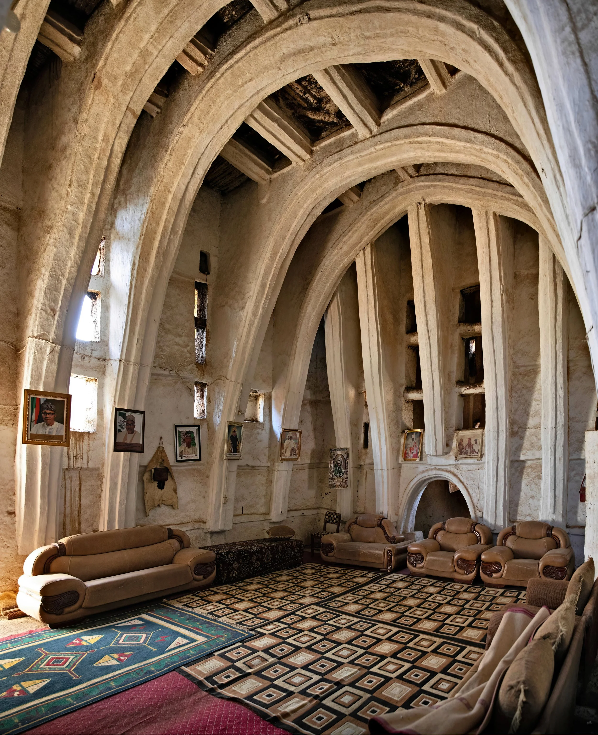 Interior of a historic building featuring high arched ceilings, beige sofas arranged along the walls, framed photographs hanging on the walls, patterned rugs on the floor, and small windows letting in natural light.