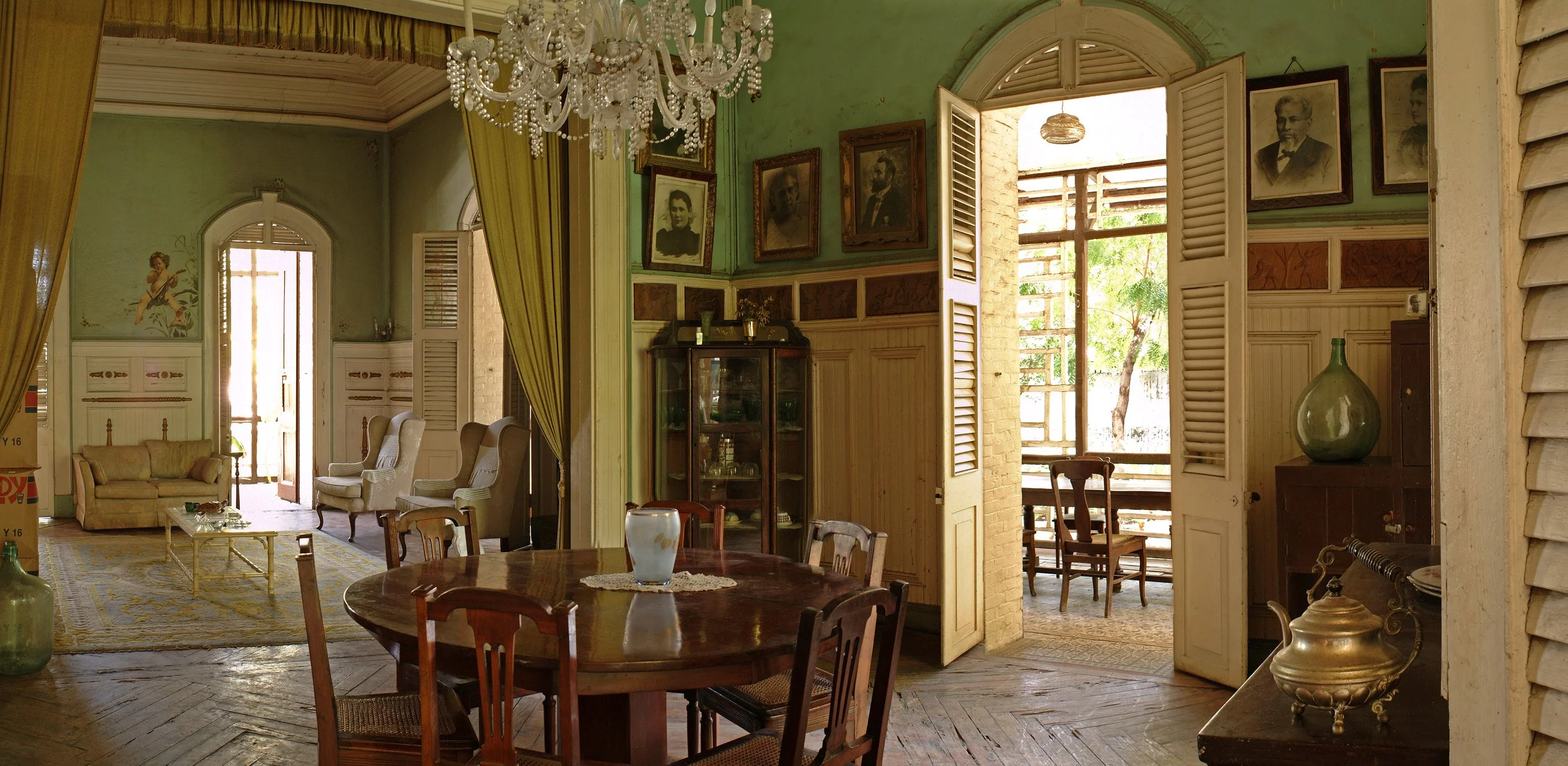 A vintage-style living and dining room with an open layout, featuring large windows with shutters, antique furniture, framed portraits on green walls, a chandelier, and a view of a balcony with trees outside.