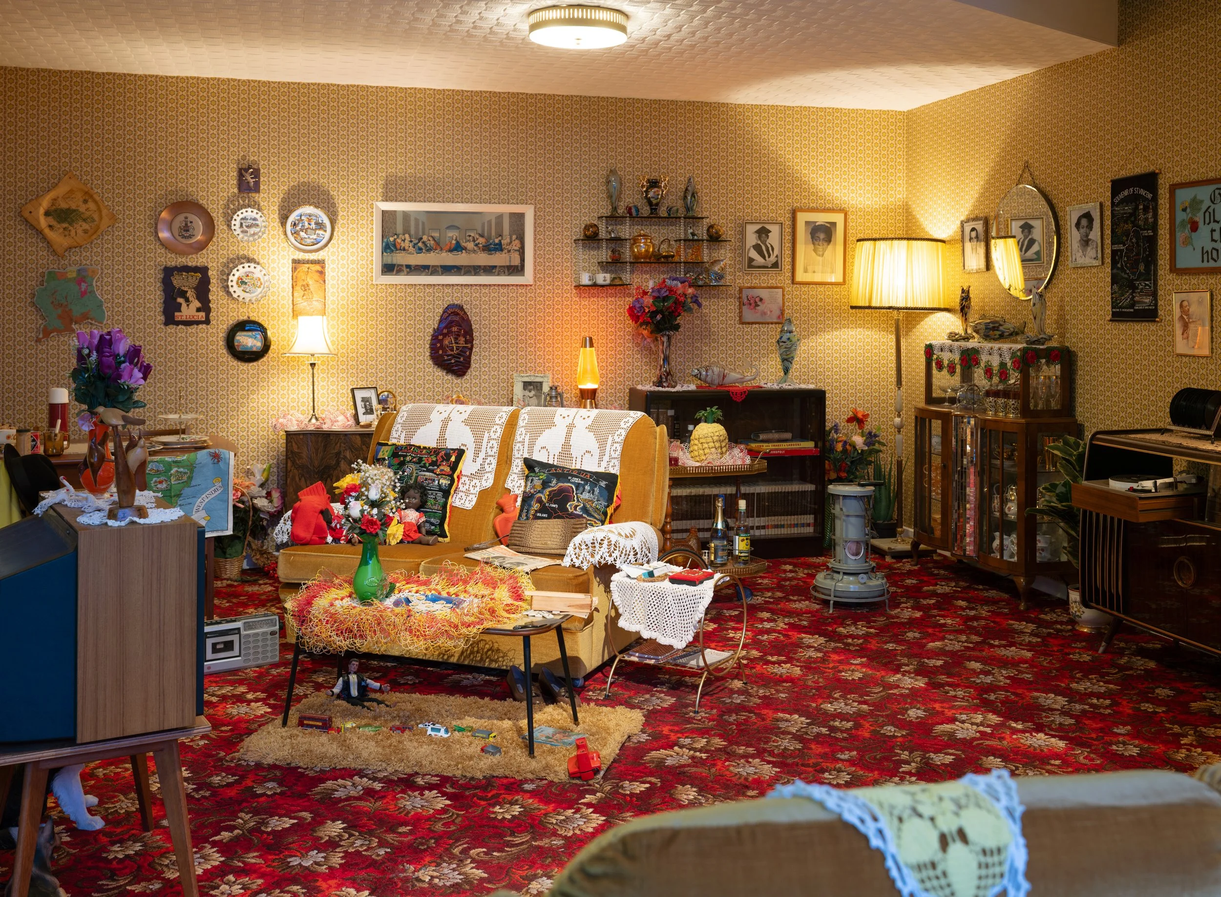 Living room with patterned wallpaper, red carpet, and various wall decorations including pictures and plates. There is a yellow sofa with embroidered covers, a table with dolls, flowers, and other decor items. A lamp, shelves with ceramics, and framed photos are also visible, along with a side table and various small objects.