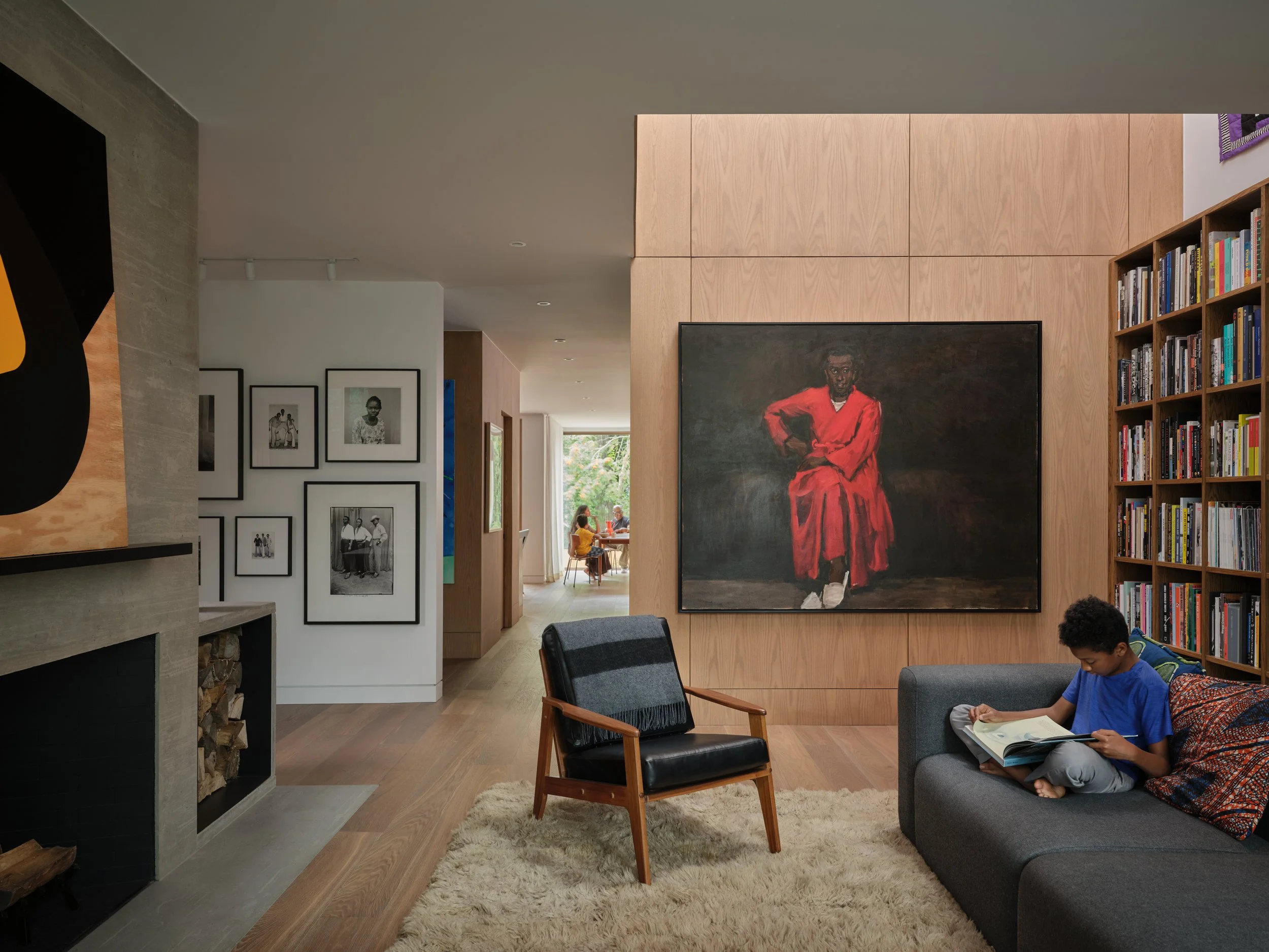 Interior view of a modern living room with wooden and white walls, a bookshelf filled with books, a large portrait painting of a person in a red outfit, black and framed photographs, and children sitting in the background.
