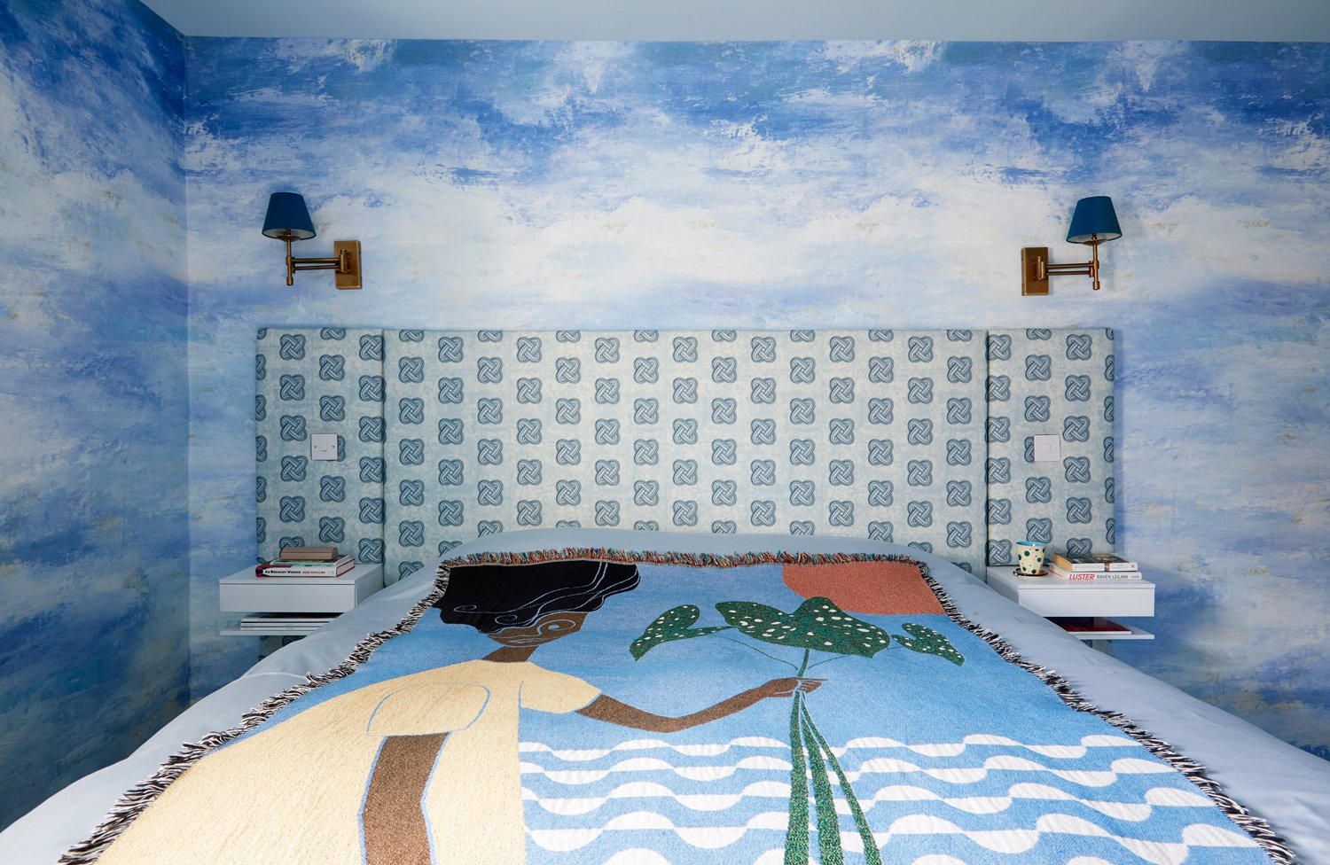 Bedroom with blue cloud-themed walls, a patterned headboard, and a colorful blanket featuring a woman holding leaves.