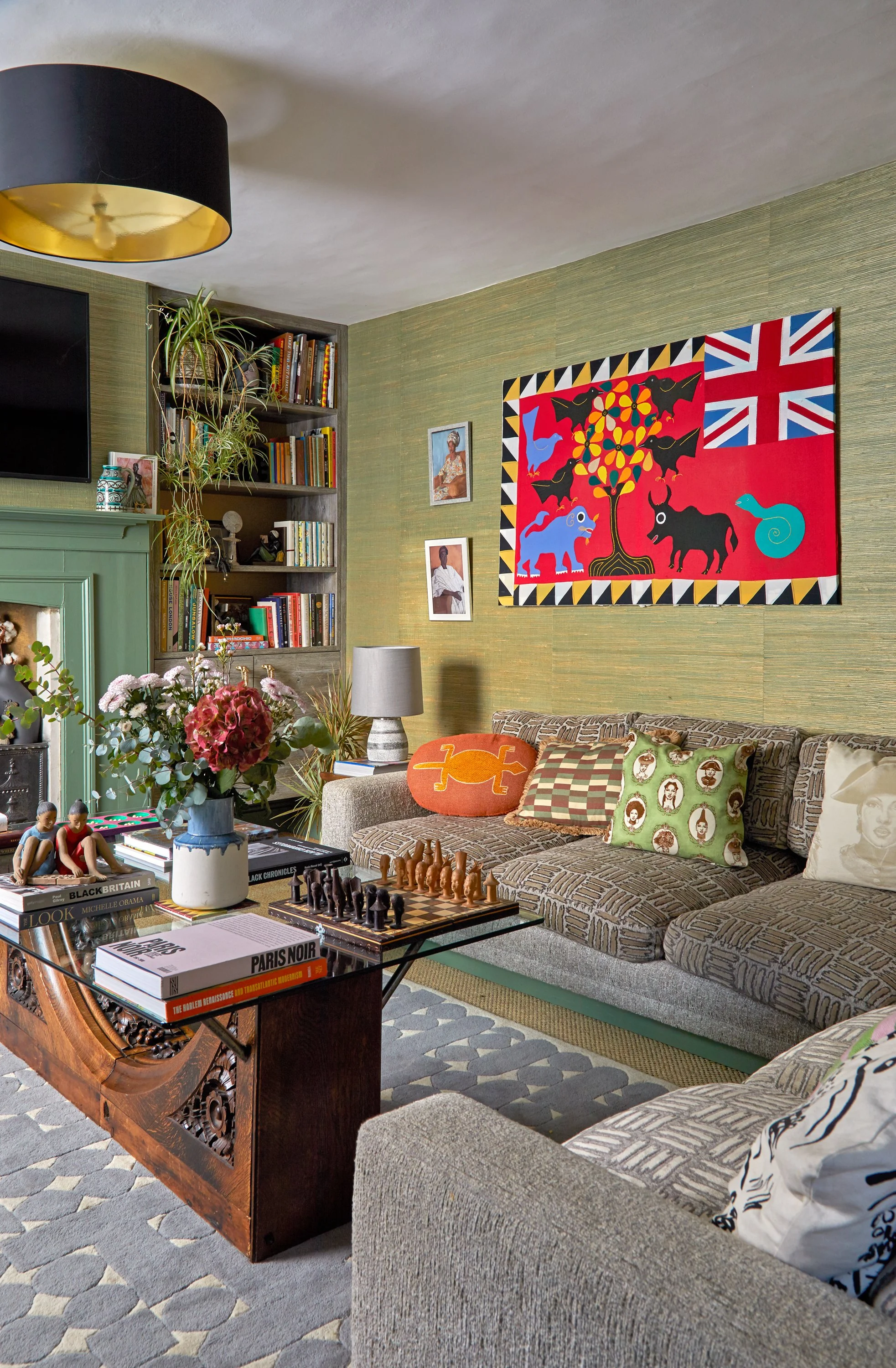 Living room with green textured wallpaper,, a bookshelf with books and plants, a TV, a beige patterned sofa with decorative pillows, a coffee table with books and a chess set, a gray lamp, and a vase of pink and white flowers on a wooden table.