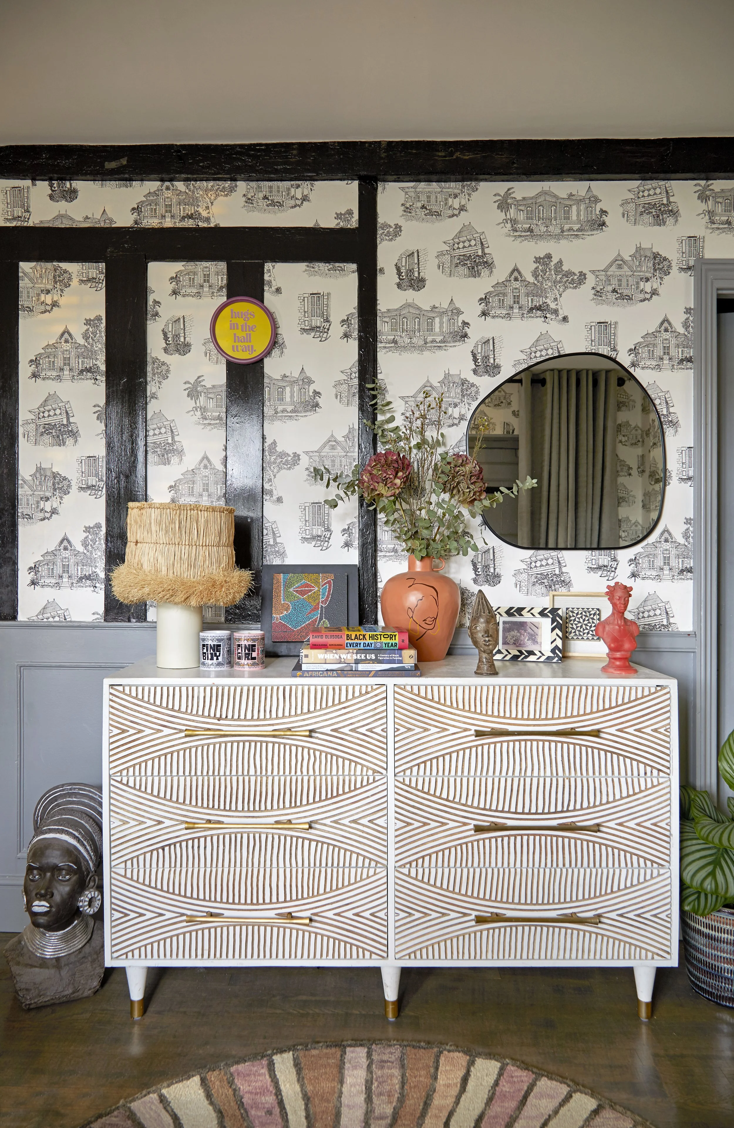A decorated interior room with a patterned light wood dresser holding art, books, and decorative objects, against a wallpapered wall with houses and trees, featuring a round mirror and a vase with flowers.