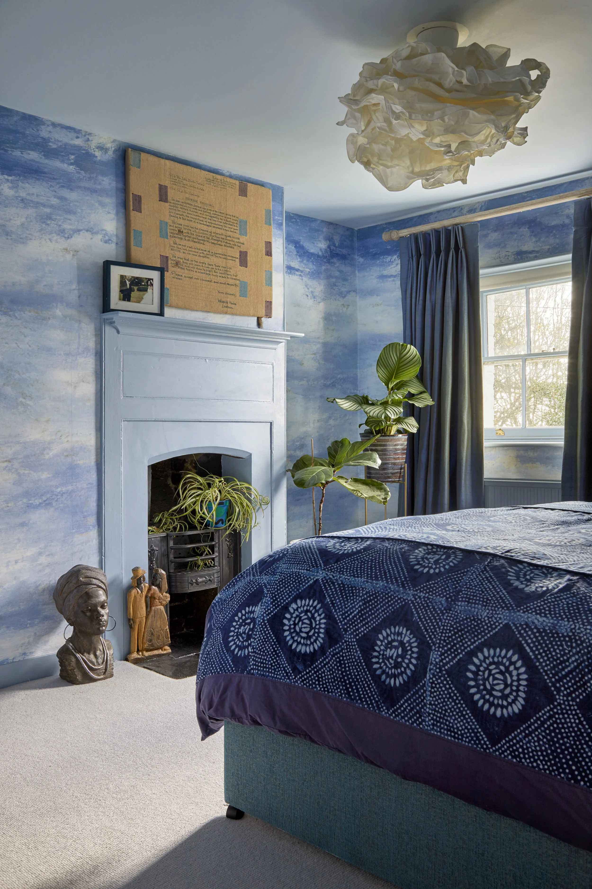 A bedroom with blue walls, a white fireplace, and a window with blue curtains. There are green plants near the fireplace, a blue and purple bedspread, and various decorative items including a bust sculpture and framed photo.