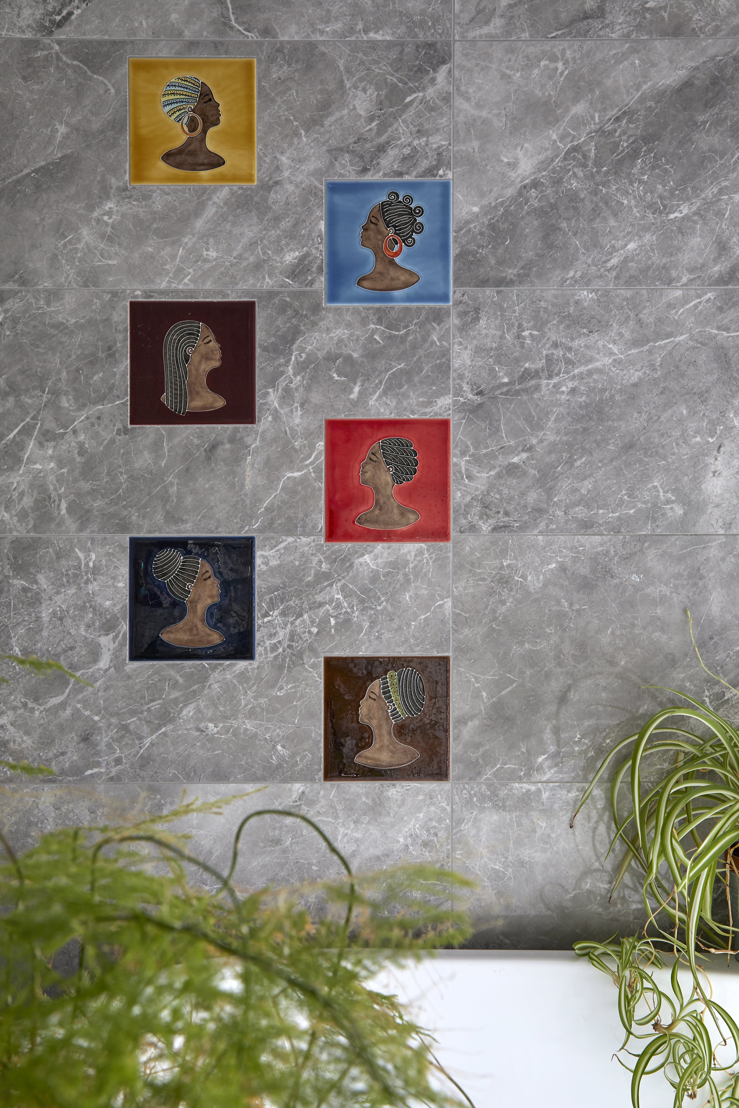 Six framed portrait artwork of women with various hairstyles and accessories, hung on a gray marble wall, partially obscured by green plants at the bottom of the image.