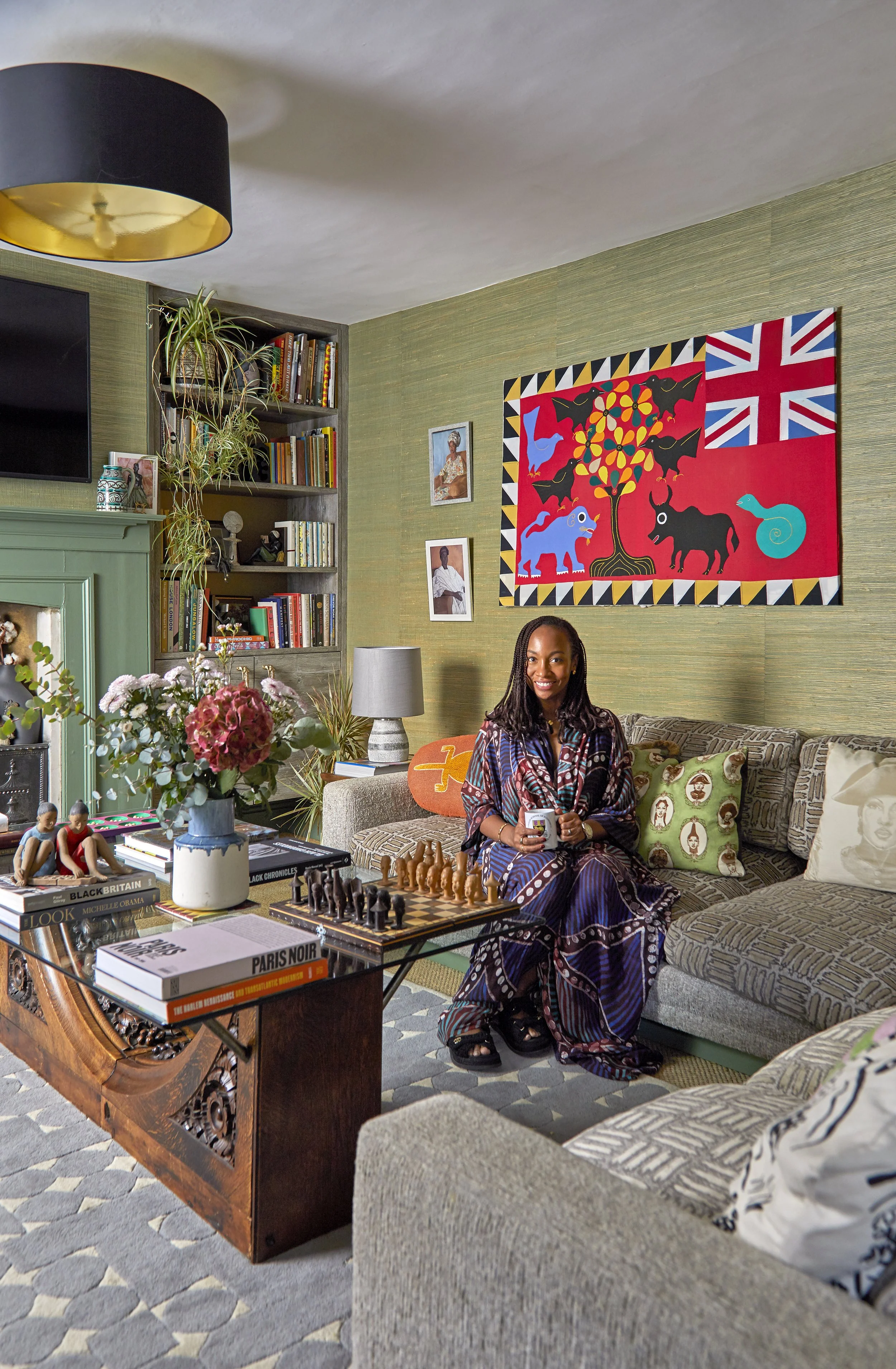 Interior view of a  living room with a woman sitting on a patterned beige sofa, smiling. The room has a green textured wall decorated with African-inspired artwork, a bookshelf filled with books, and various decorative items. A dark fireplace mantel, a lamp, a vase with pink flowers, and a chess set on a wooden table are also visible. The floor has a patterned rug, and there are additional decorative pillows on the sofa.