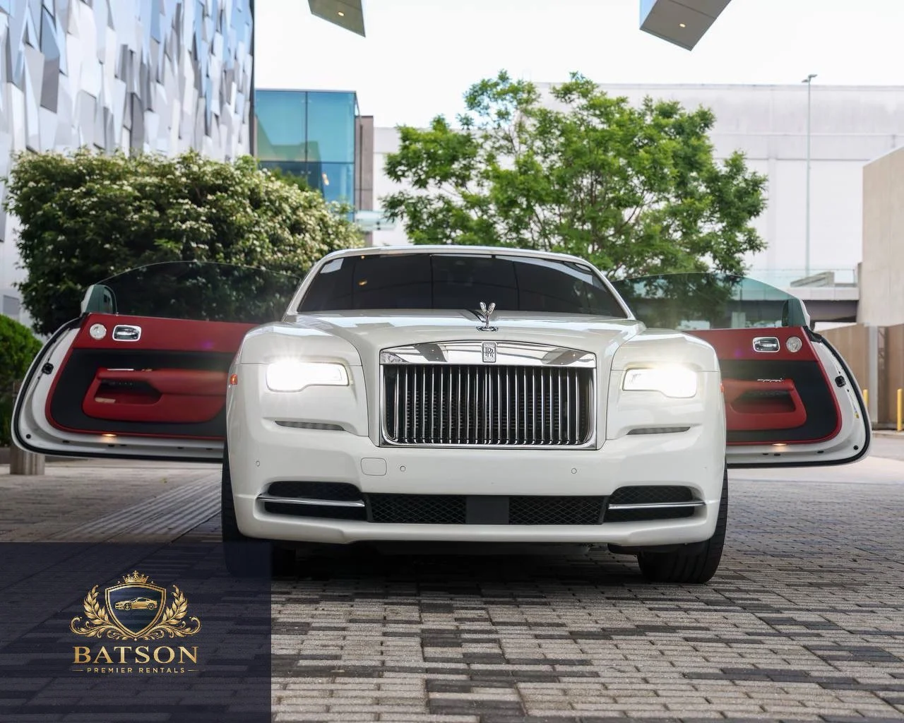A white luxury Rolls Royce car with open doors, parked in an urban area with modern buildings, trees, and clear skies in the background.