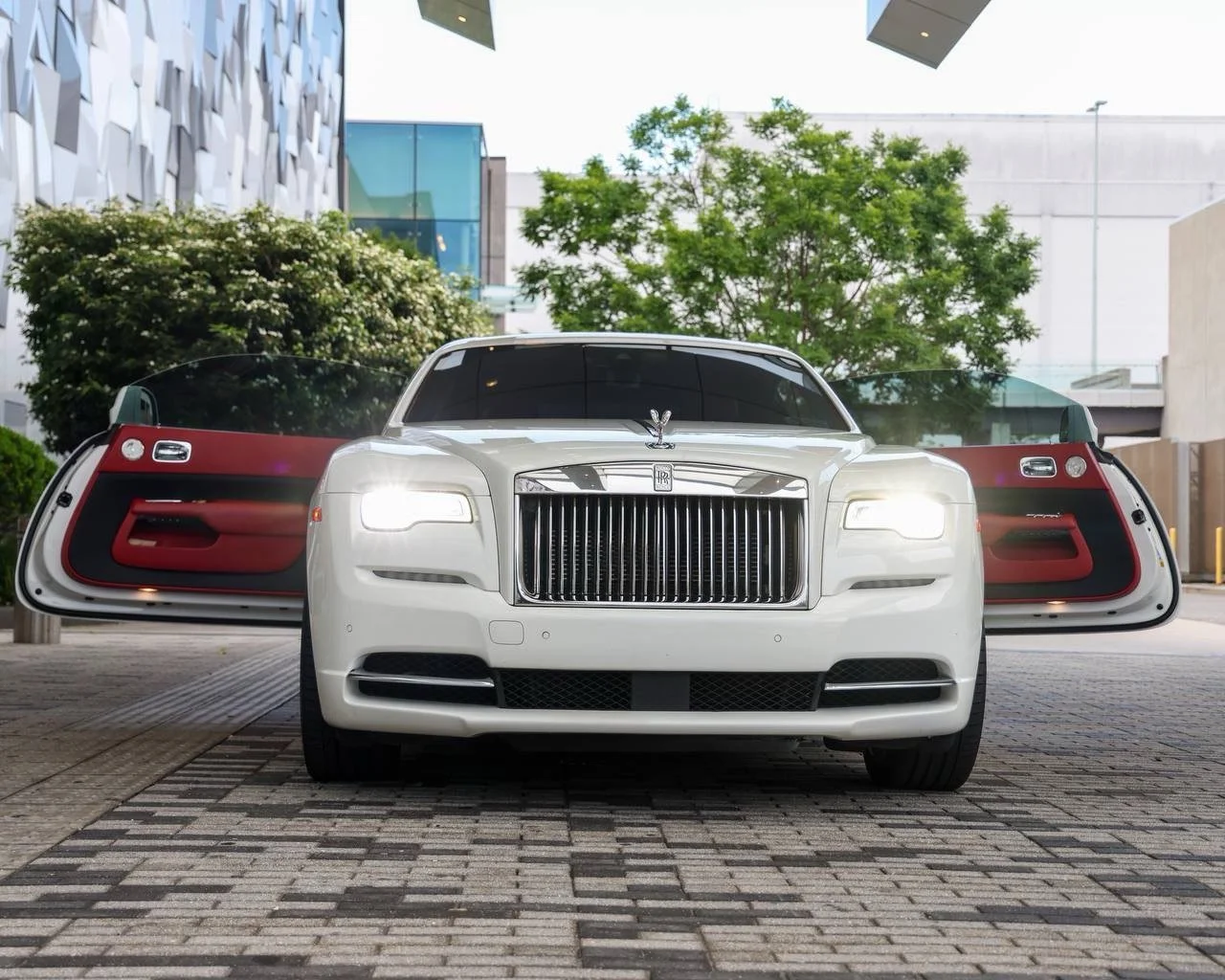 A white luxury car, Rolls-Royce, with open driver and passenger doors, parked outdoors in an urban area with modern buildings, trees, and a brick pavement outside in Charlotte, North Carolina.