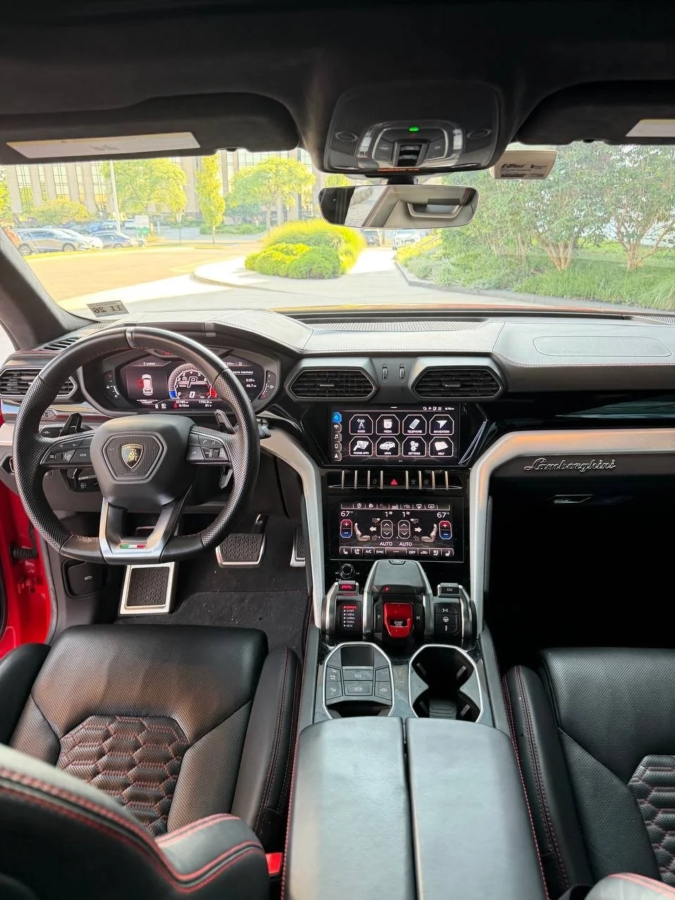 Interior of a luxury Lamborghini sports car with a digital dashboard, steering wheel, and center console with controls and touchscreen display, parked in a landscaped area.