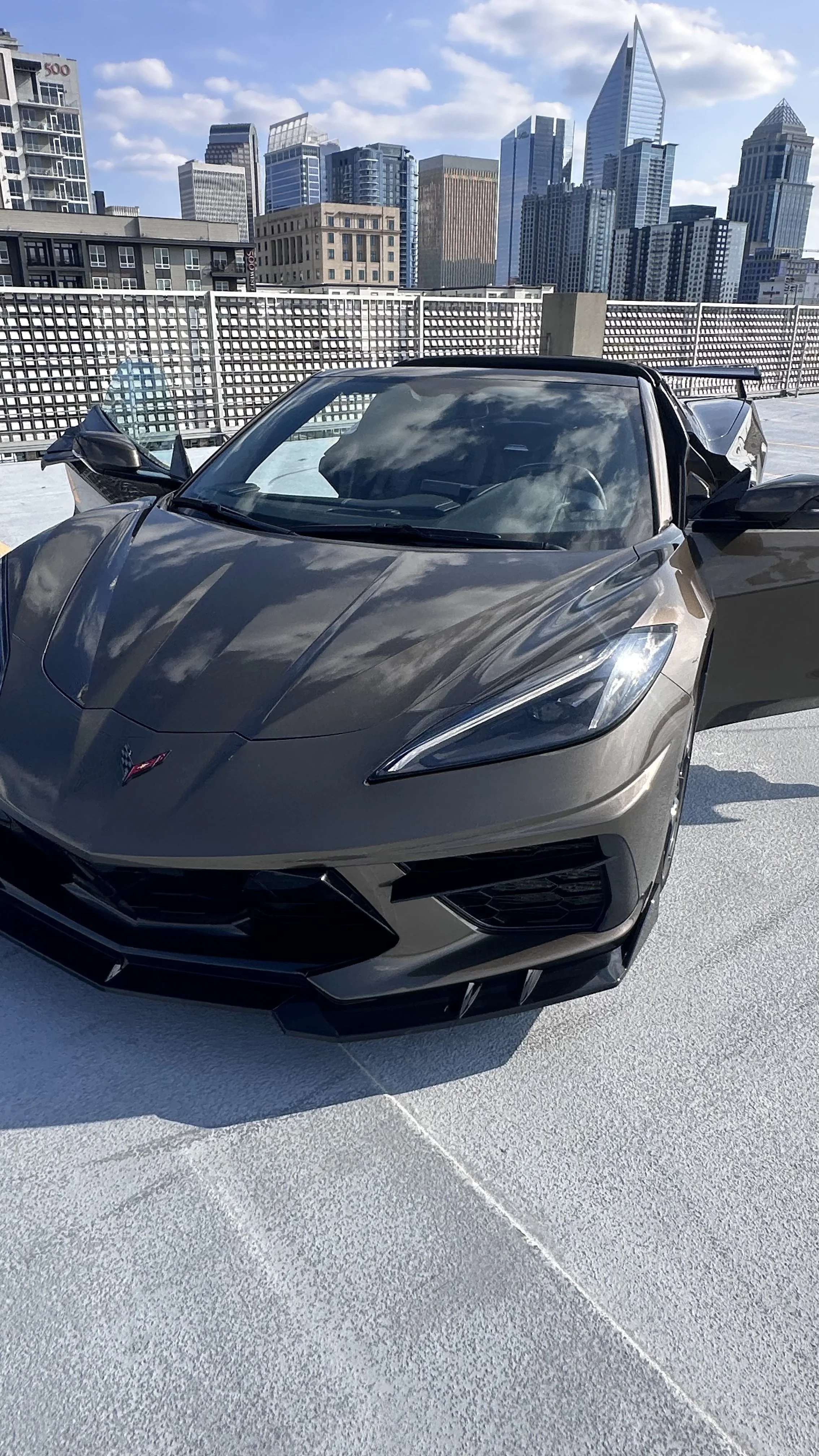 A Bronze Chevrolet Corvette C8 sports car parked on a rooftop parking lot In Charlotte, North Carolina with a city skyline in the background.
