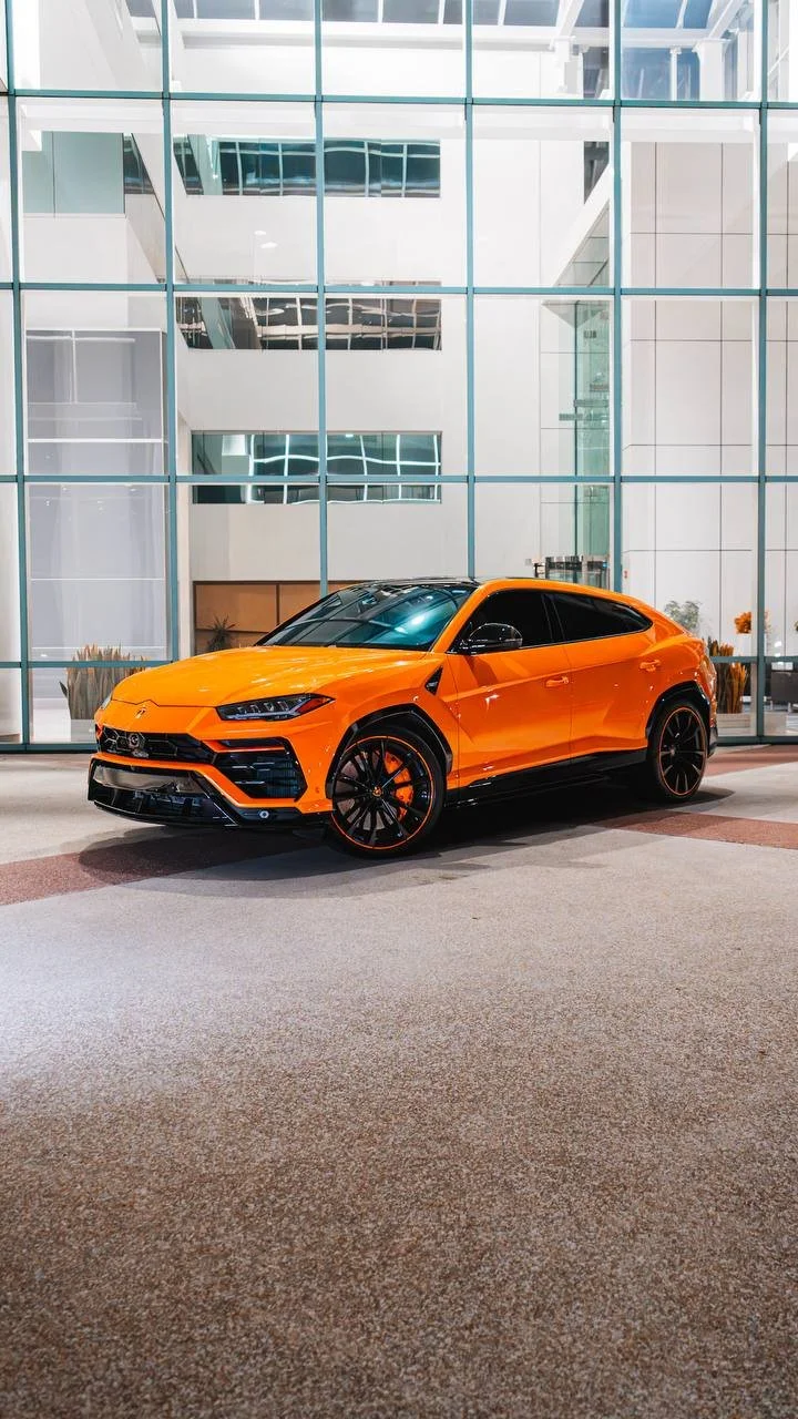 An orange Lamborghini SUV parked in front of a modern glass building in Charlotte, North Carolina at night.