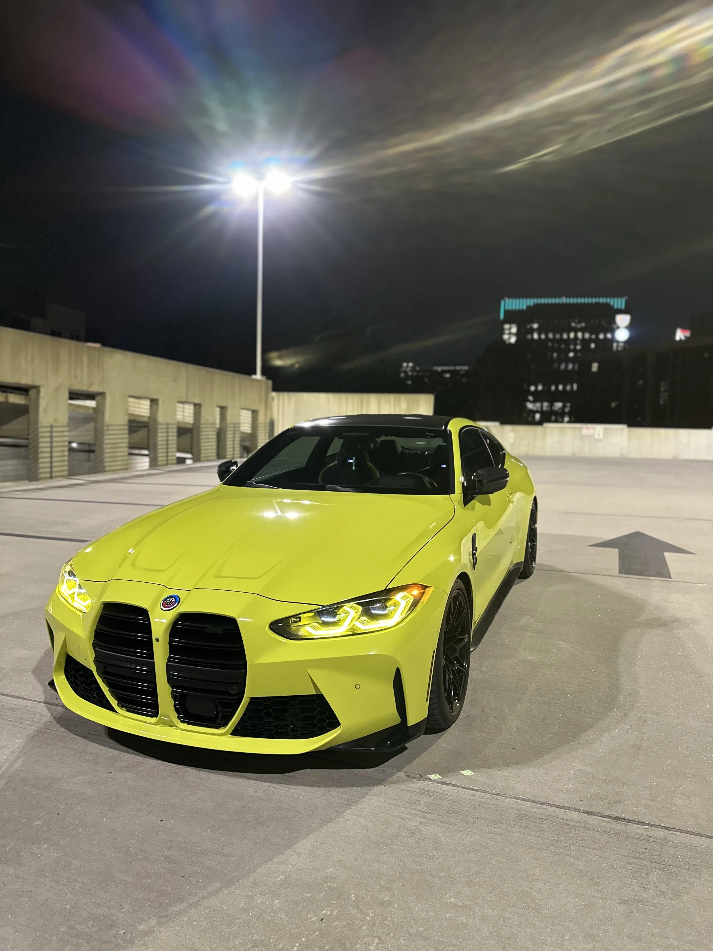 Bright yellow BMW M3 and M4 sports car parked in a multi-level parking garage at night with city buildings in the background in Charlotte, North Carolina