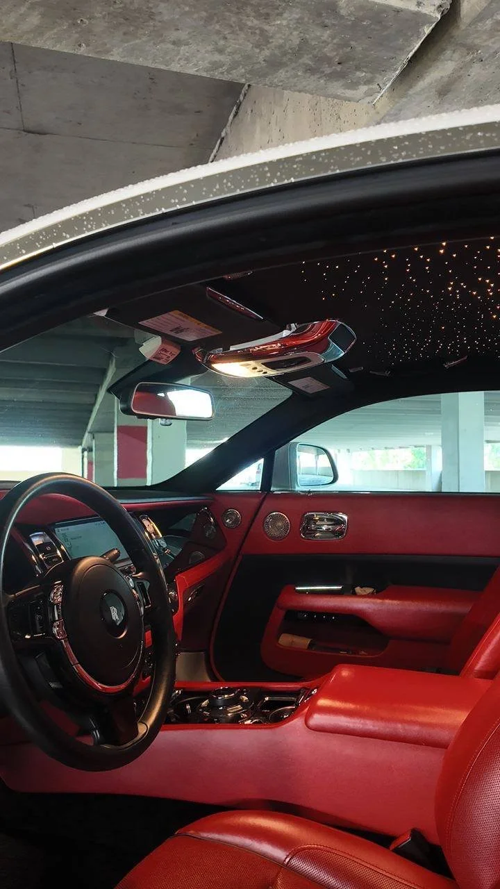Interior of a luxury car with red leather seats, a black steering wheel, a starry ceiling lighting feature, and a view of a parking garage exterior.