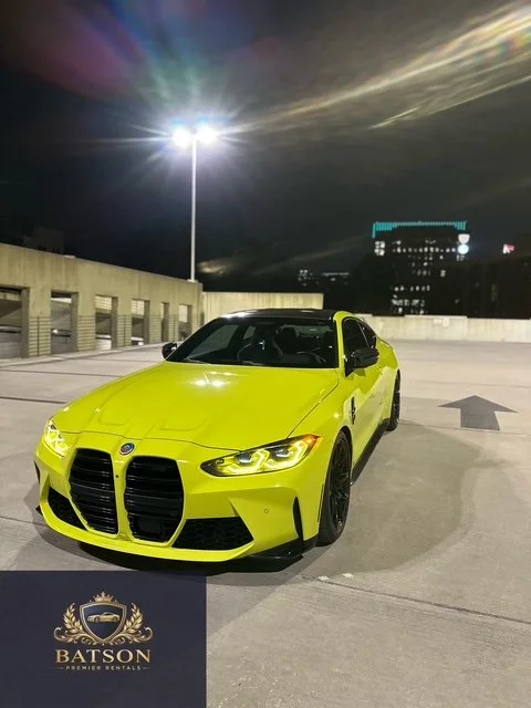 A bright yellow M4 COMP car parked on a rooftop parking lot at night with city buildings and a street light in the background.