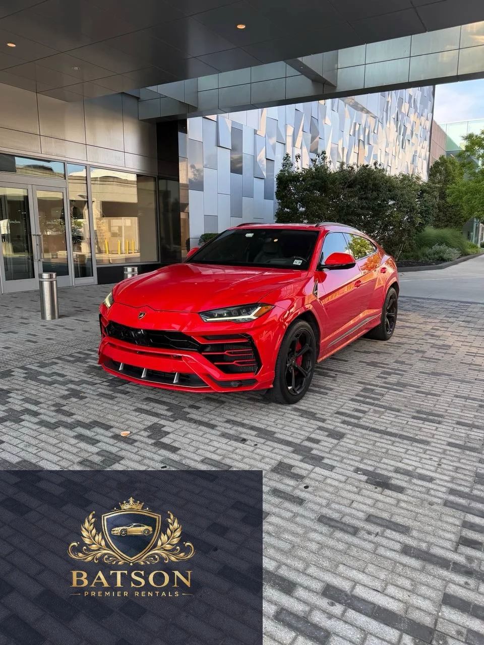 Red Lamborghini Urus parked outside a modern building with glass doors and windows, surrounded by trees and greenery.