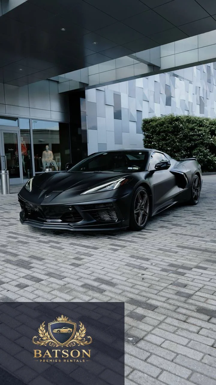A matte black C8 Corvette parked outside a modern building with a glass entrance and decorative metal wall panels, near a small bush.