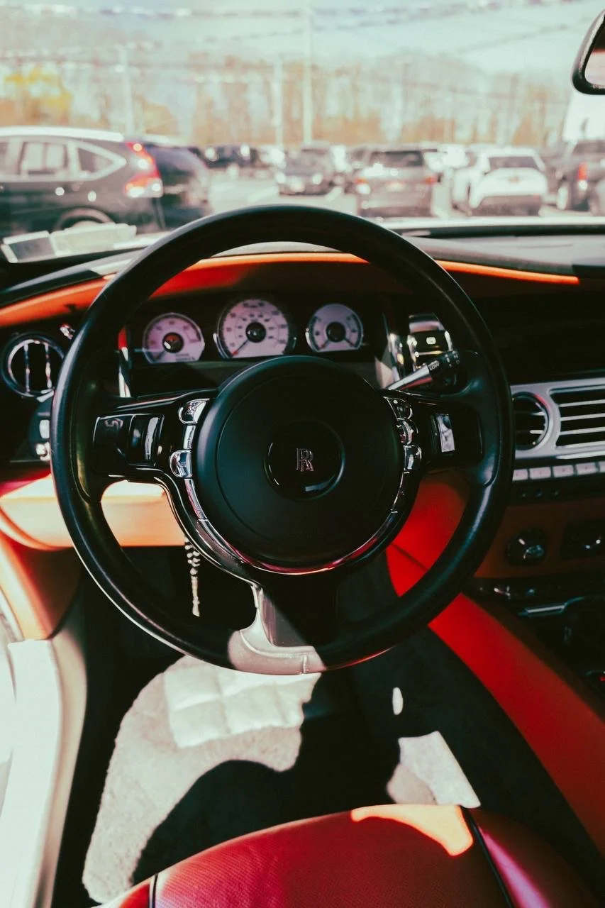 Interior view of a luxury car showing the steering wheel with a Rolls-Royce emblem, dashboard with gauges, and parking lot seen through the windshield.