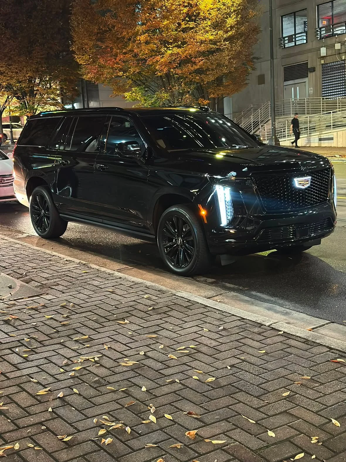 A black Cadillac SUV parked on a city street at night with autumn trees in the background and a person walking on the sidewalk.