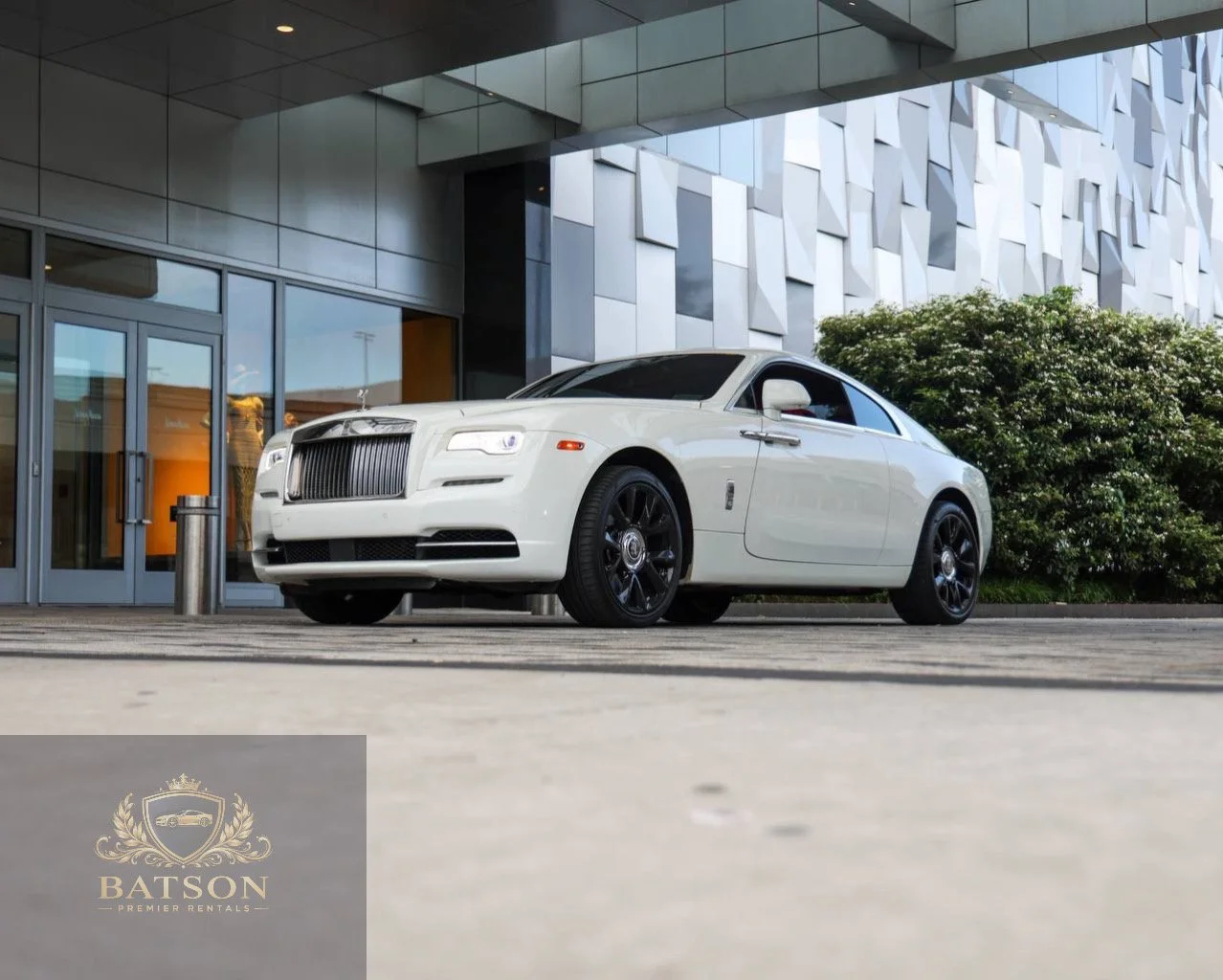 A white Rolls-Royce Wraith, parked in front of a modern building with glass doors and a patterned facade, with greenery nearby, during daytime.
