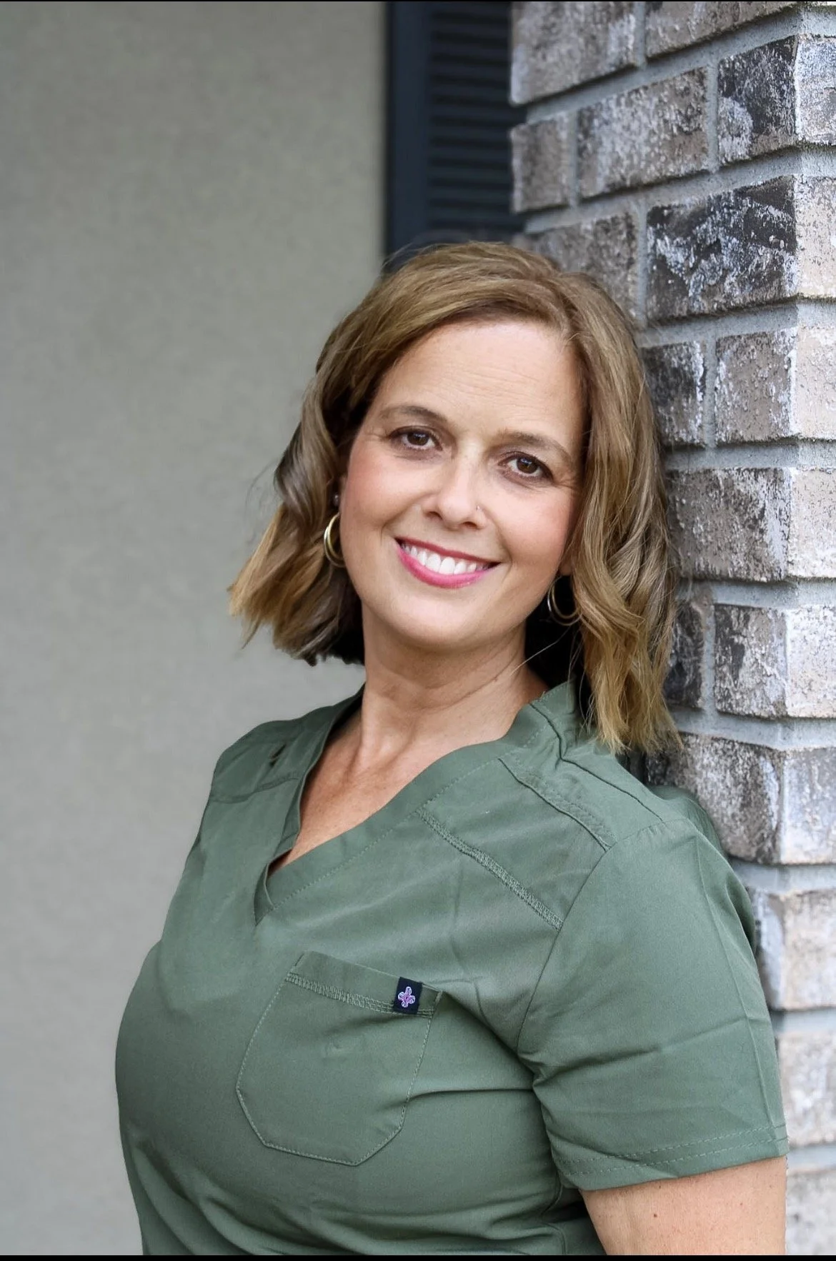 A woman with shoulder-length brown hair, wearing hoop earrings and a green medical scrub top, smiling while leaning against a brick wall with a neutral-colored background.