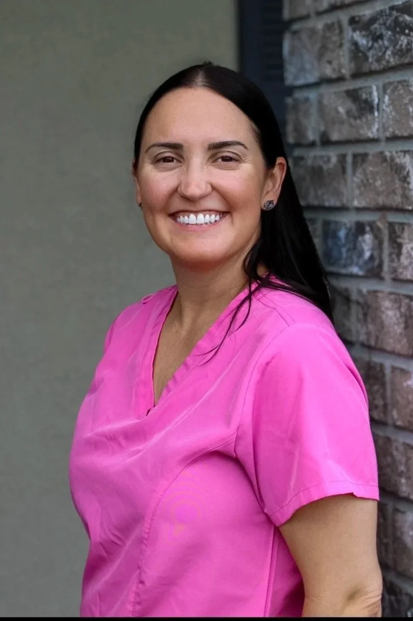 A woman with long dark hair, smiling, wearing pink scrubs, standing outdoors near a brick wall.