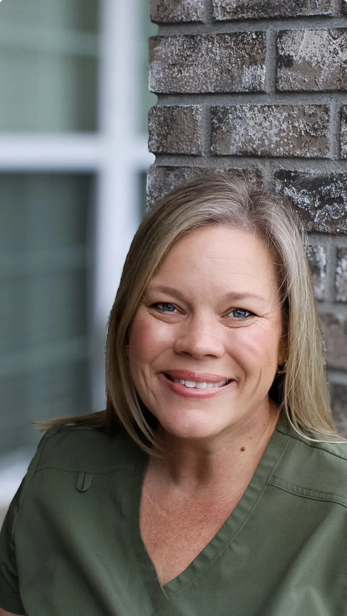 Close-up of a smiling woman with blonde hair, blue eyes, wearing an olive green shirt, standing outdoors against a brick wall and window.
