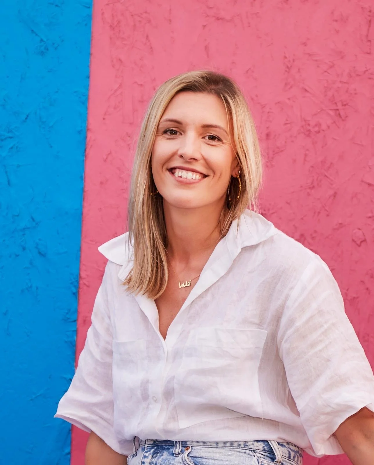A smiling woman with shoulder-length blonde hair wearing a white shirt, gold hoop earrings, and a necklace with an Arabic name, standing in front of a multicolored wall with blue and pink sections.