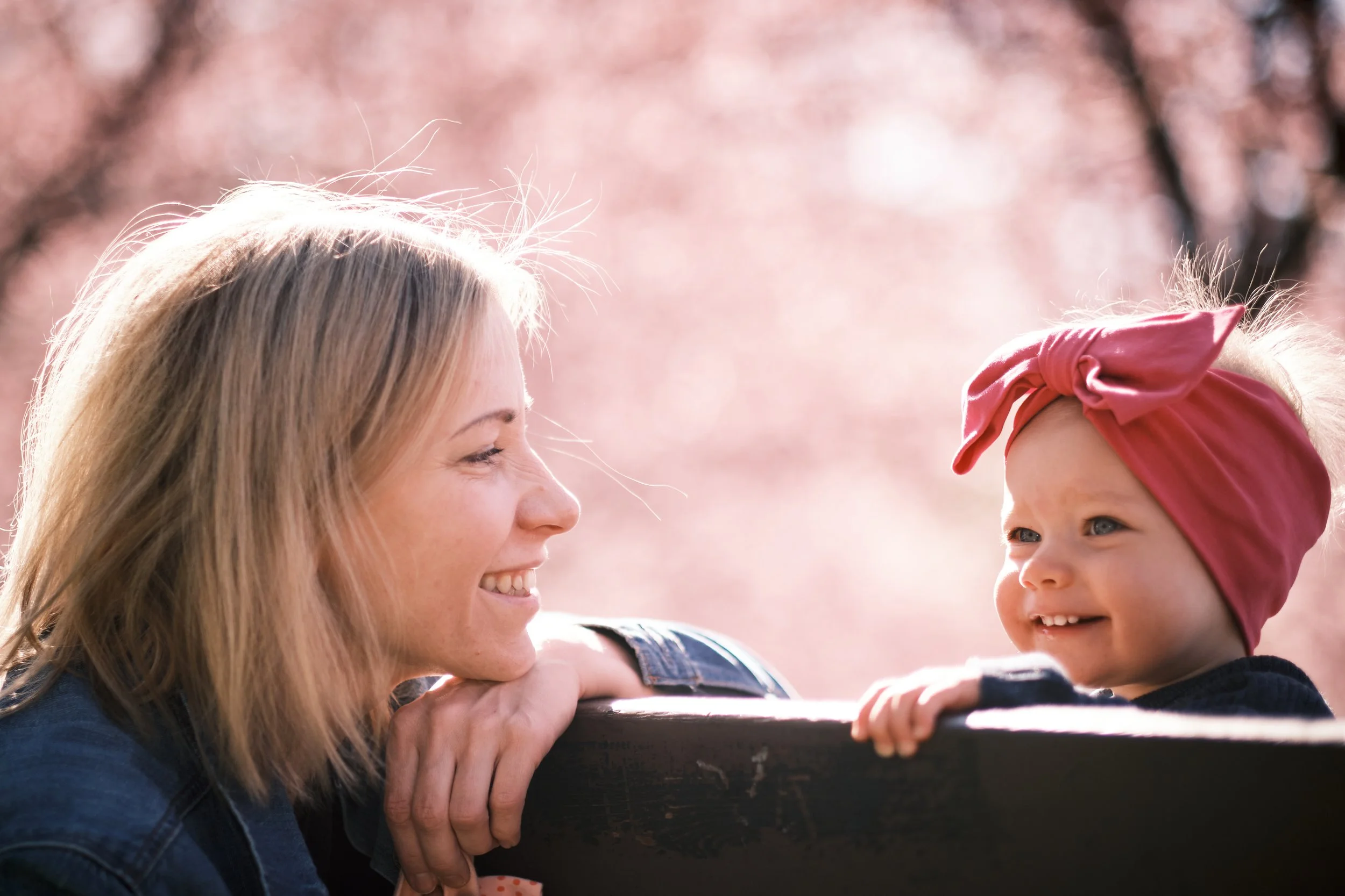 A woman and a young girl smiling at each other outdoors on a sunny day, with the woman leaning on a wooden surface. The girl is wearing a pink headband with a bow, and the background features pink cherry blossoms.