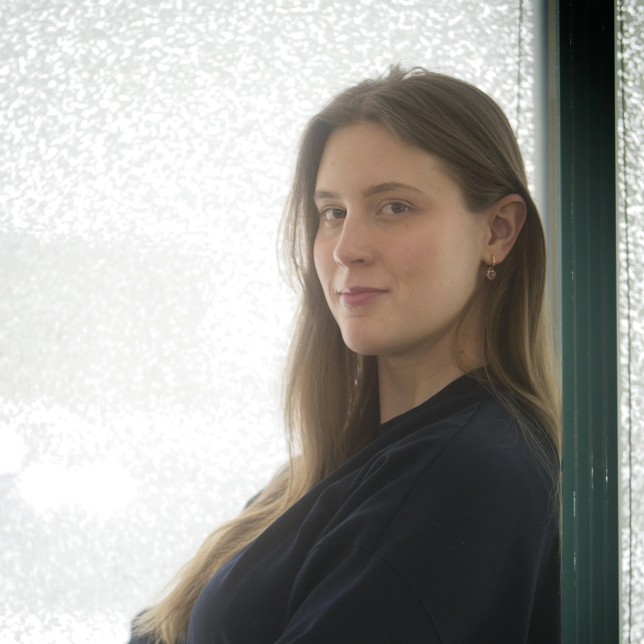 A woman with long brown hair, wearing earrings and a black top, standing near a window with frosted glass, looking at the camera with a slight smile.