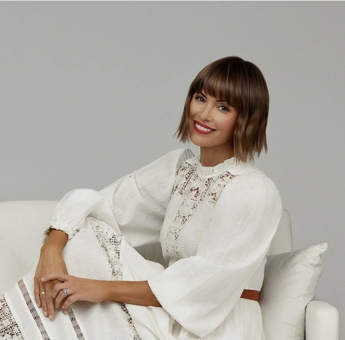 A woman with short brown hair and red lipstick sitting on a white sofa, smiling, wearing a white lace dress against a gray background.