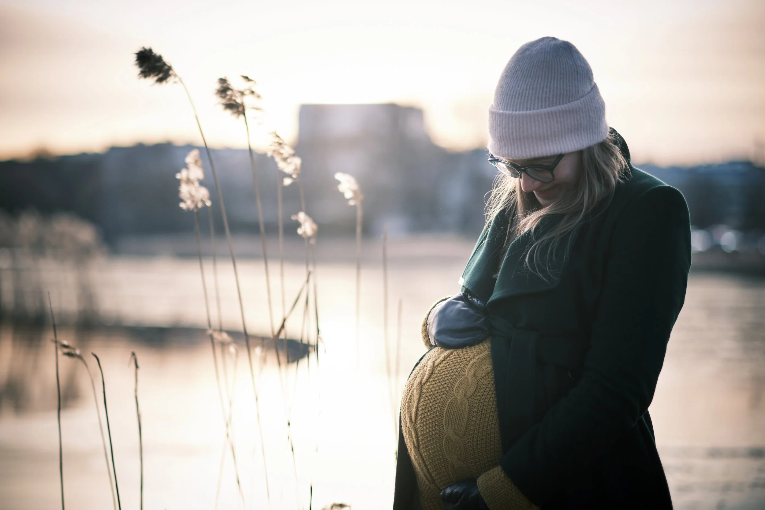 A woman wearing glasses, a white knit hat, and dark coat holding a pregnant woman or a mannequin wearing a yellow sweater by a body of water at sunset.