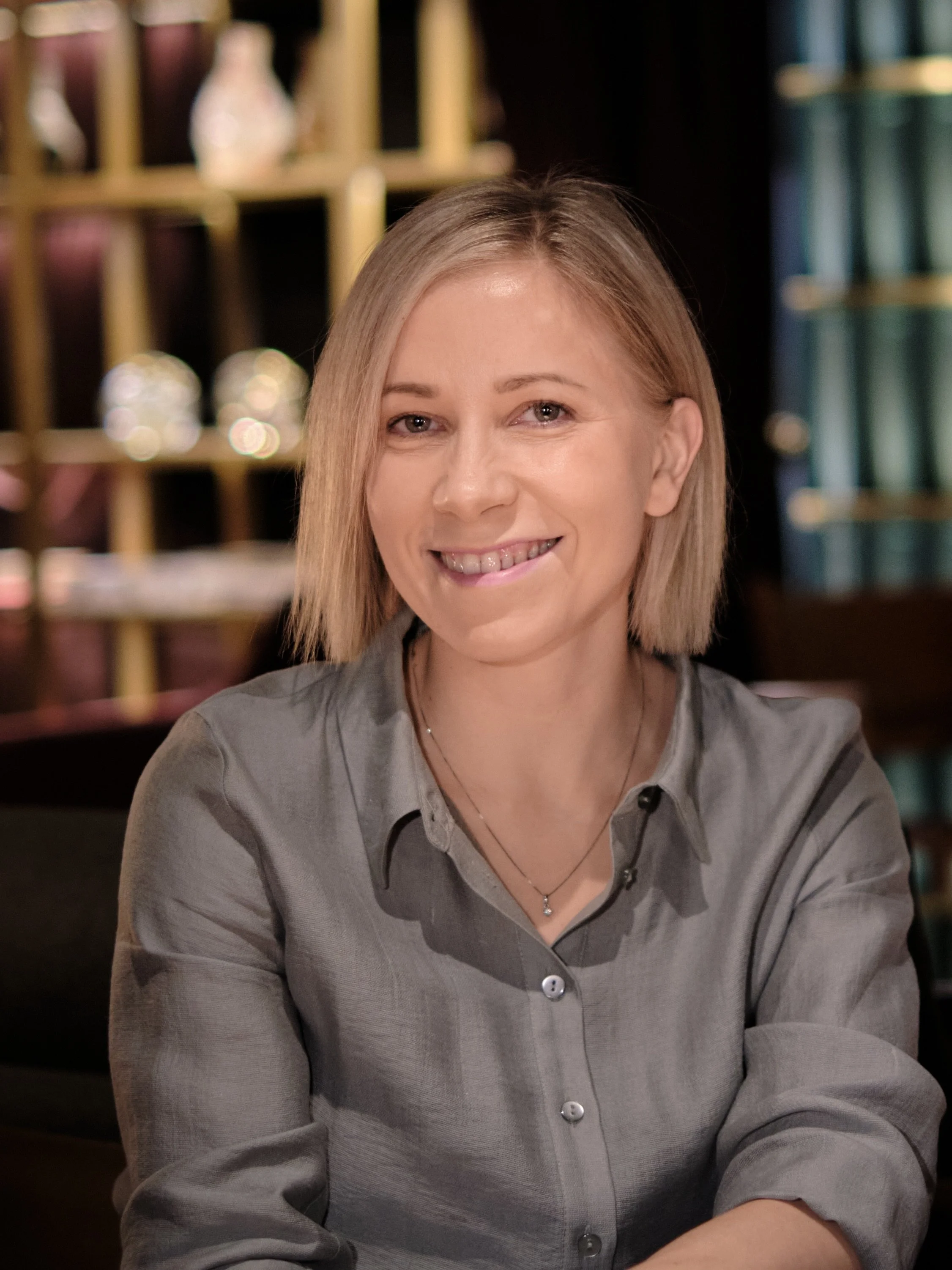 A woman with shoulder-length blonde hair, smiling and wearing a gray button-up shirt, sitting indoors in front of shelves filled with decorative items and books.