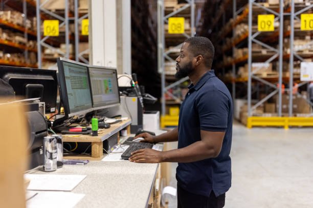 A man working in a warehouse, standing at a desk with two computer monitors, surrounded by shelves filled with boxes and supplies.