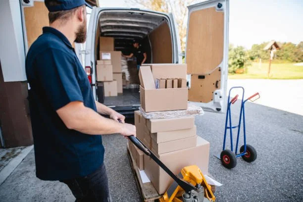Two men are handling packaging and moving boxes from a white delivery van with an open back door, with one man using a hand truck and the other inside the van.