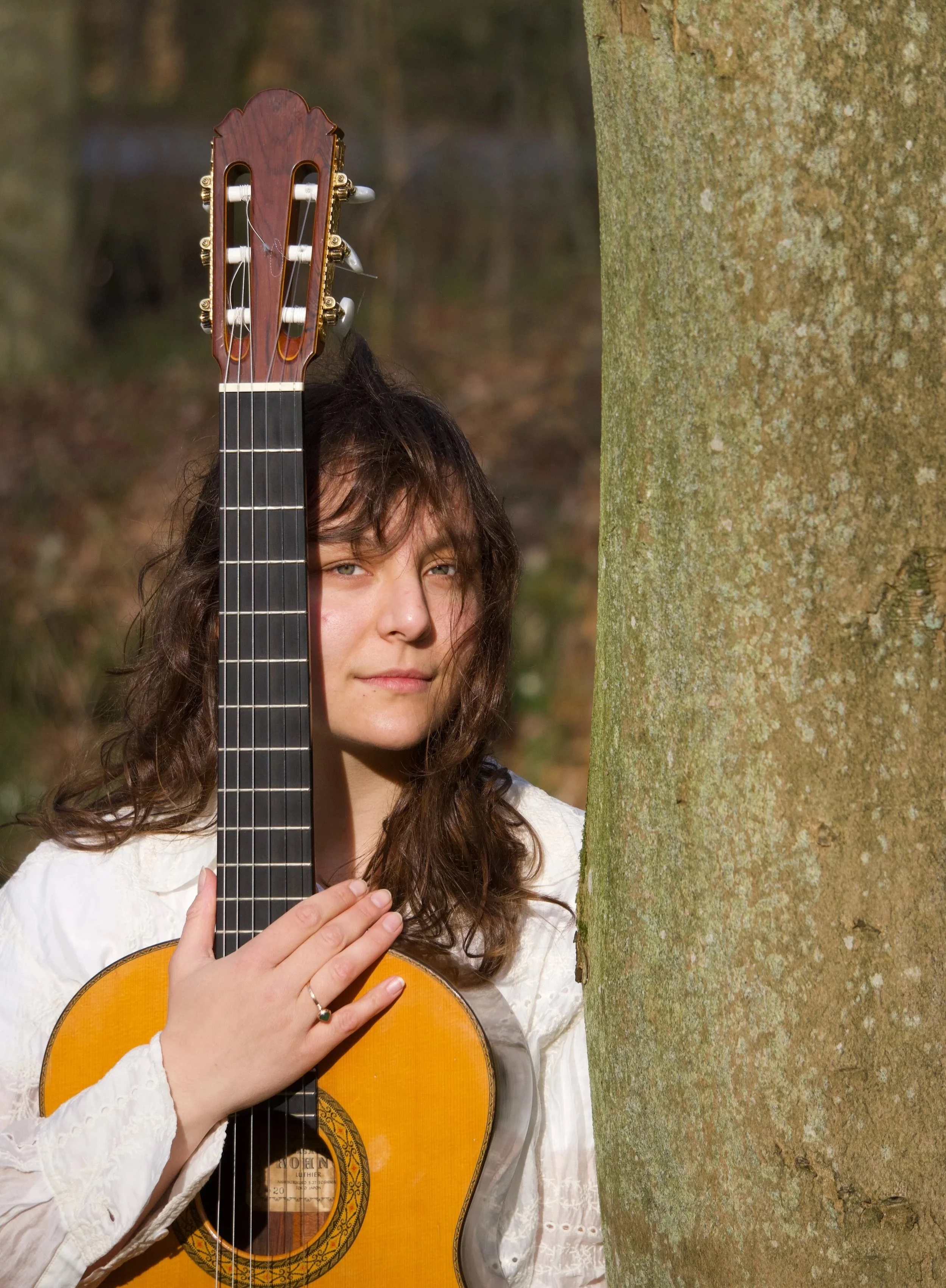 A young woman with brown curly hair holding an acoustic guitar partially behind a tree trunk in a wooded outdoor setting.