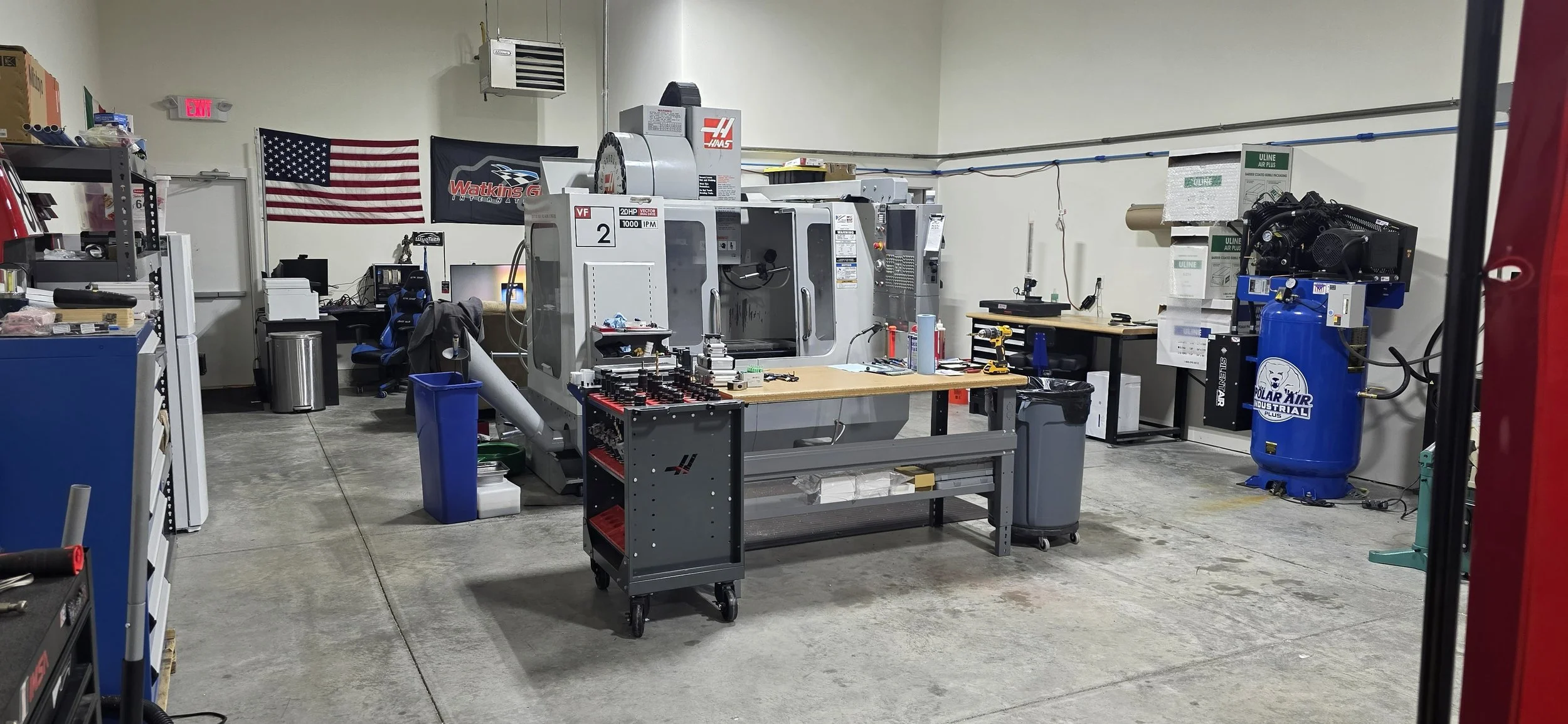 Workshop with industrial machines, tools, and workstations, including a large CNC machine, workbenches with tools, and storage shelves, with American flags on the wall.
