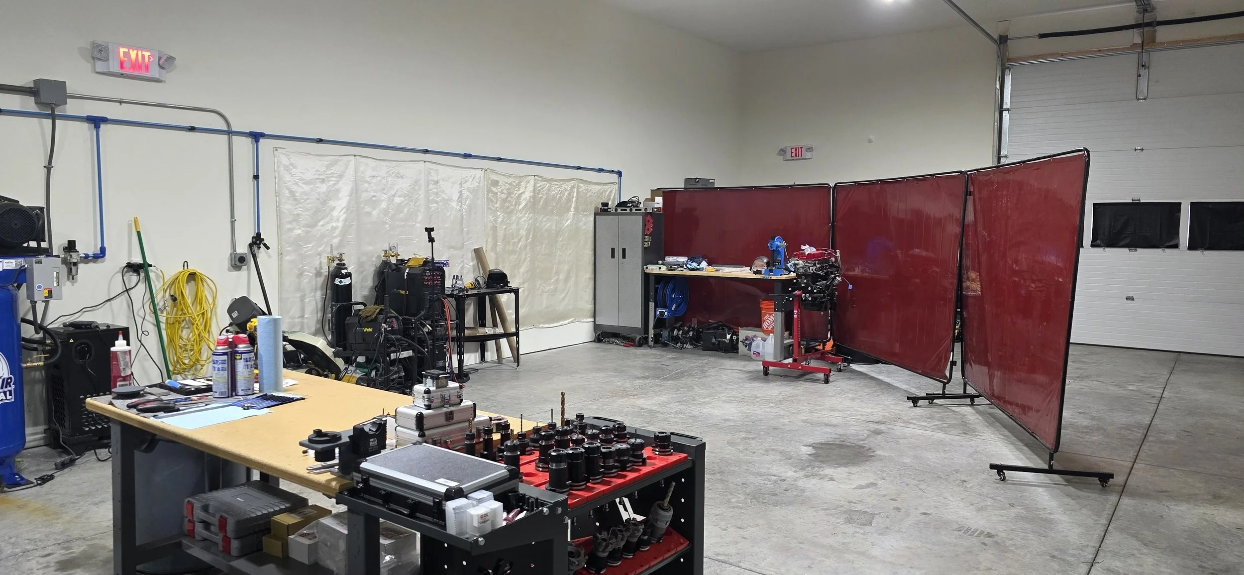 A workshop with a table covered in supplies, machinery, and tools. The background has red partitions, a white wall, and a large garage door.